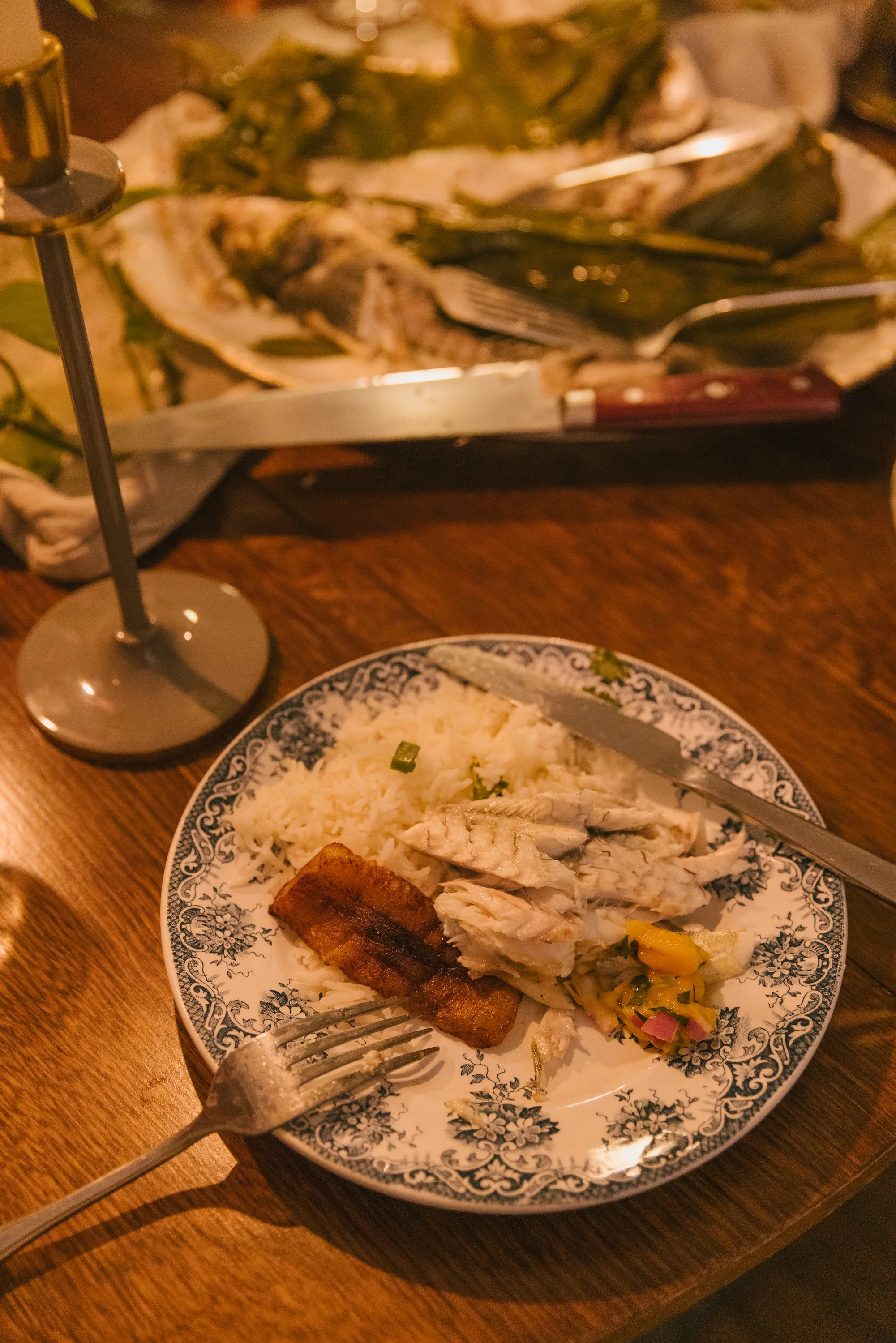 A plate of white rice with shredded chicken, a slice of pork belly, and a vegetable medley, on a decorative patterned plate. In the background, a dish with grilled fish, a fork, and a wine glass with a dark beverage are visible.