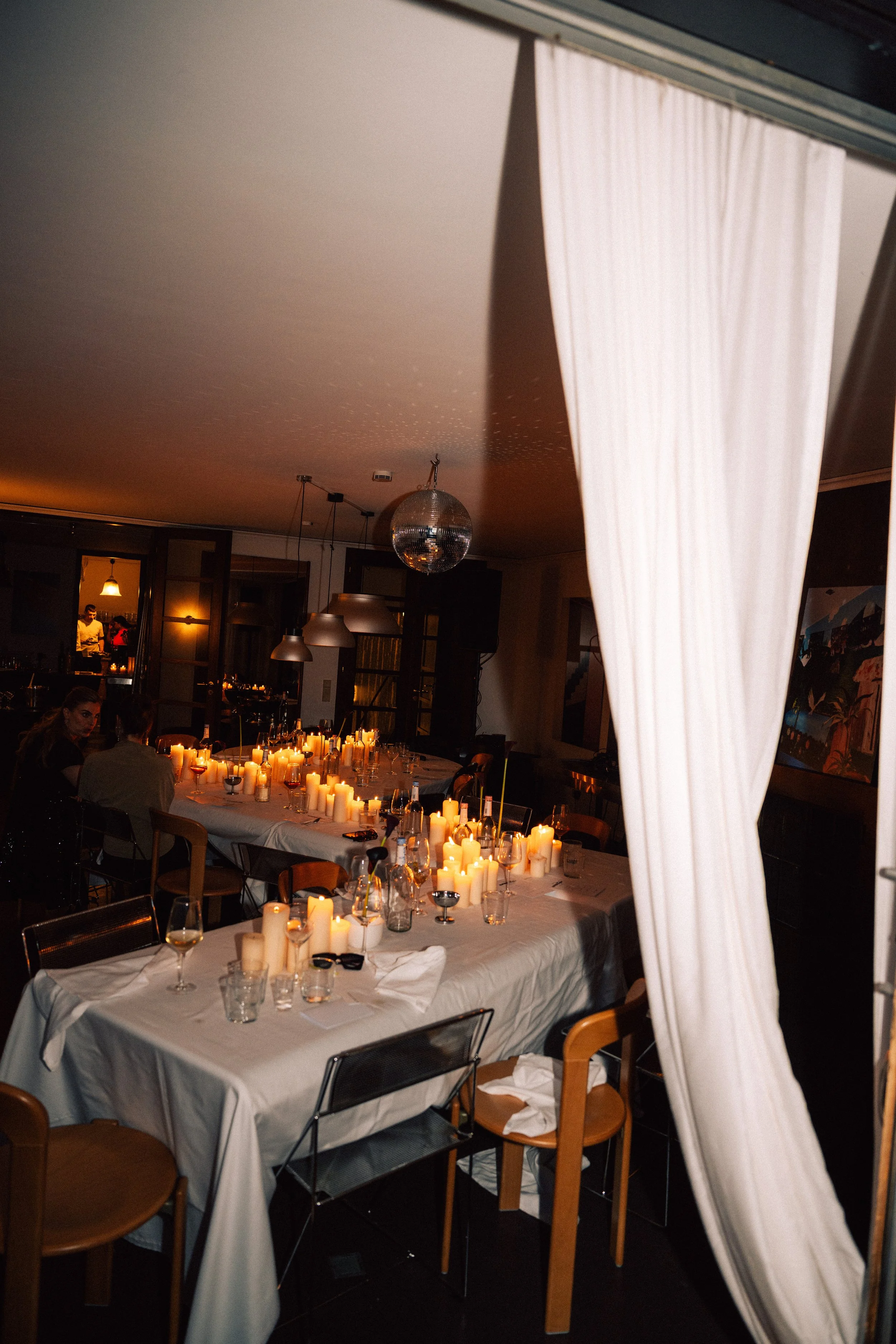 Dinner party with a long table decorated with numerous candles, wine glasses, and utensils; people seated and engaged in conversations; dim lighting and framed artwork on the walls, with a disco ball hanging from the ceiling.