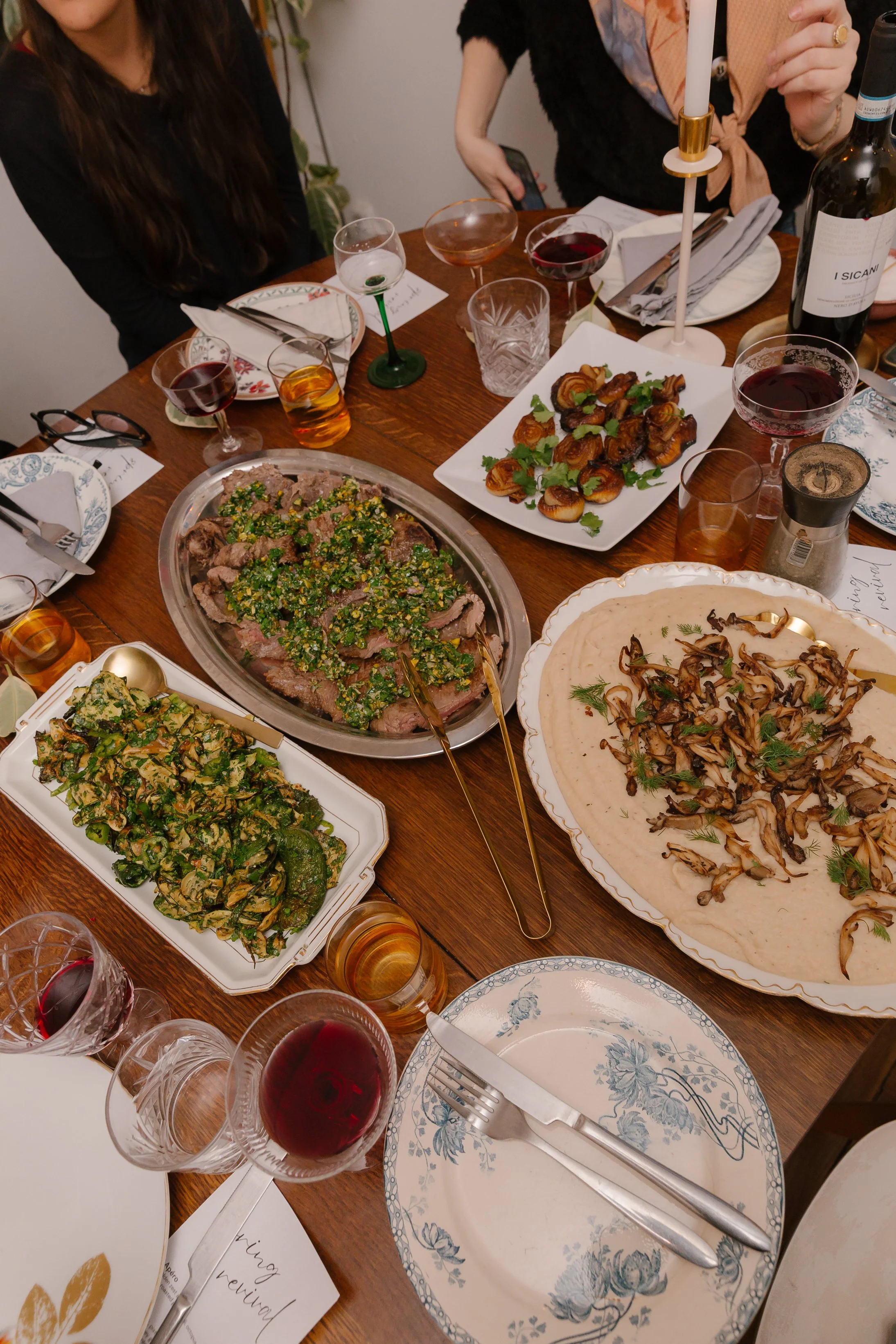 A dinner table with various dishes including sliced beef with herbs, roasted vegetables, and a flatbread with mushrooms. There are glasses of red wine, a candle, and dinnerware set for multiple people, with some people sitting around the table.