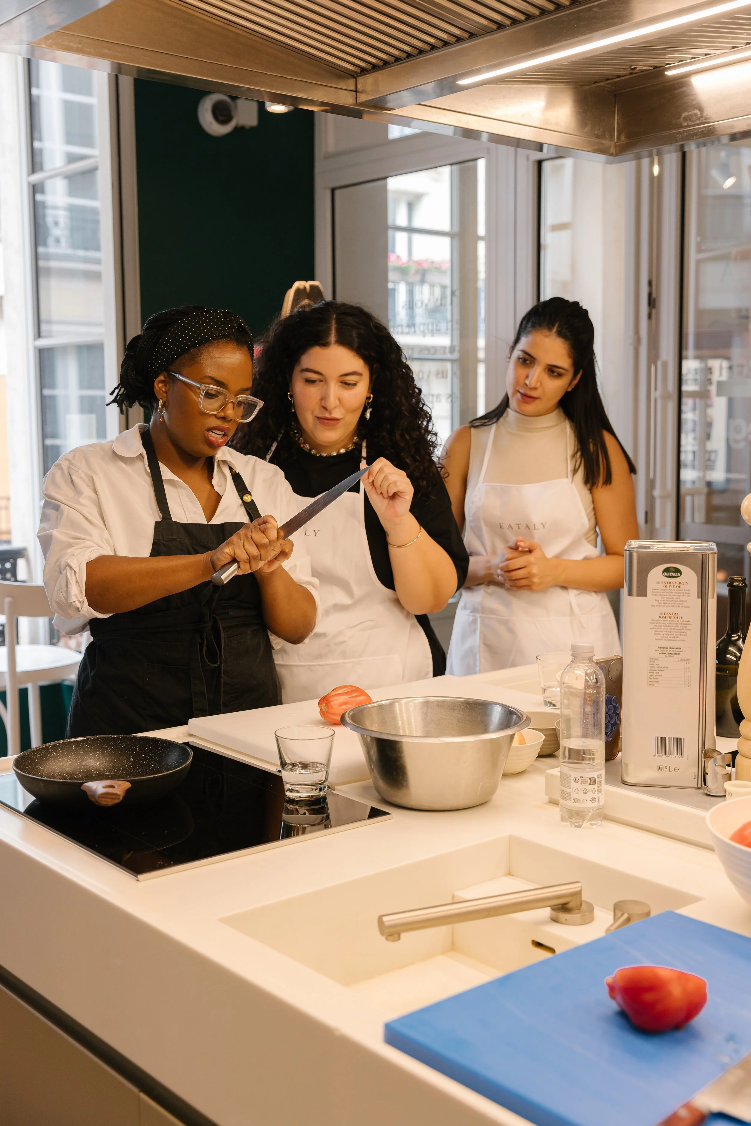 Three women in aprons participating in a cooking class in a modern kitchen with large windows. They are gathered around a kitchen island with various cooking tools and ingredients, including a knife, a tomato, and a bowl.