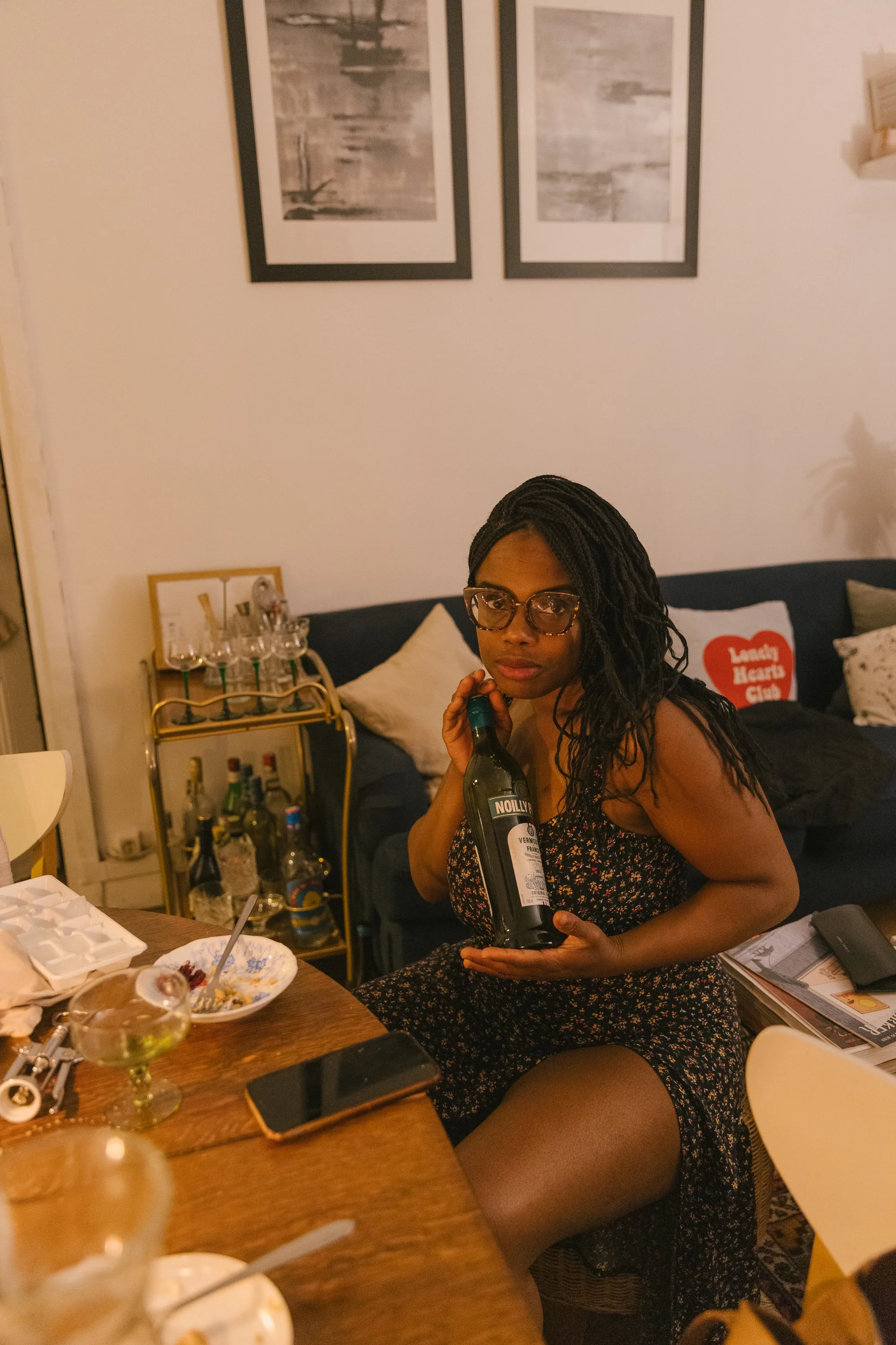 A woman with glasses and long braided hair sitting at a table holding a wine bottle in her right hand. The table has food, drinks, and miscellaneous items. Behind her is a sofa with pillows, including one with text. The wall has two framed black and 