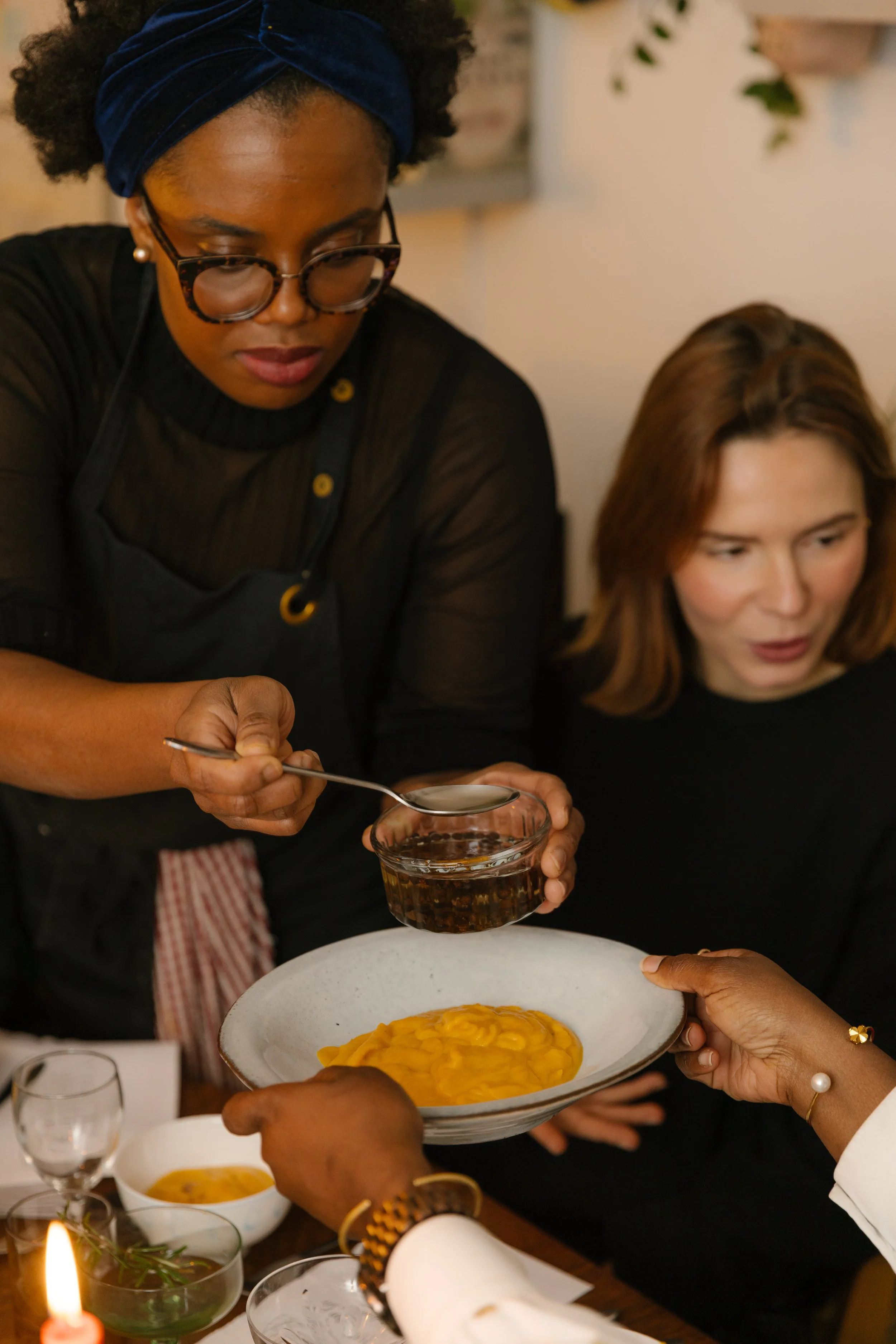 A woman wearing glasses and a headband is pouring sauce from a spoon onto a plate of pasta, with another woman in the background. The table has glasses, bowls, and candles.