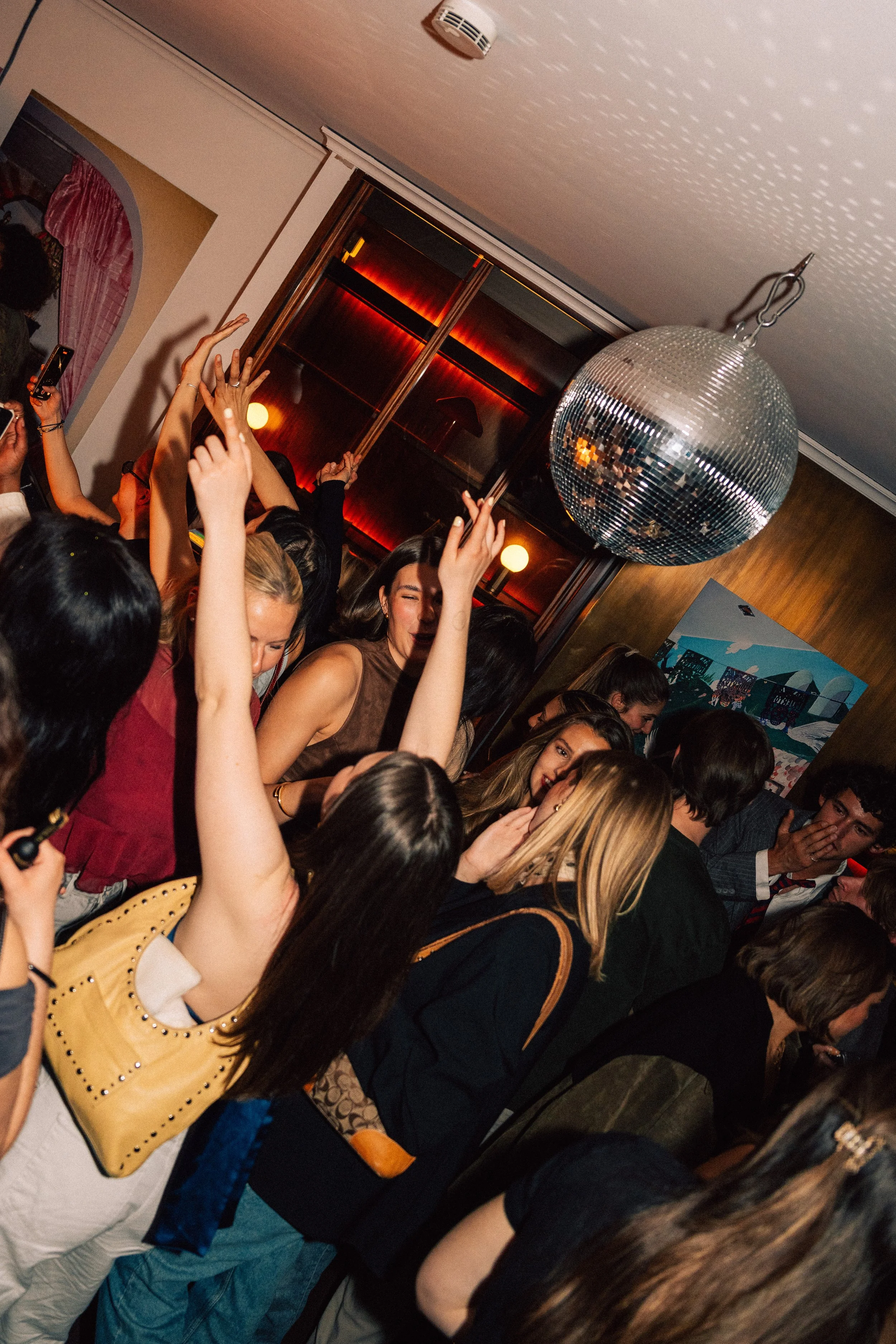 A crowded party or dance floor with people smiling, dancing, and raising their hands, illuminated by colorful lights and a disco ball hanging from the ceiling.