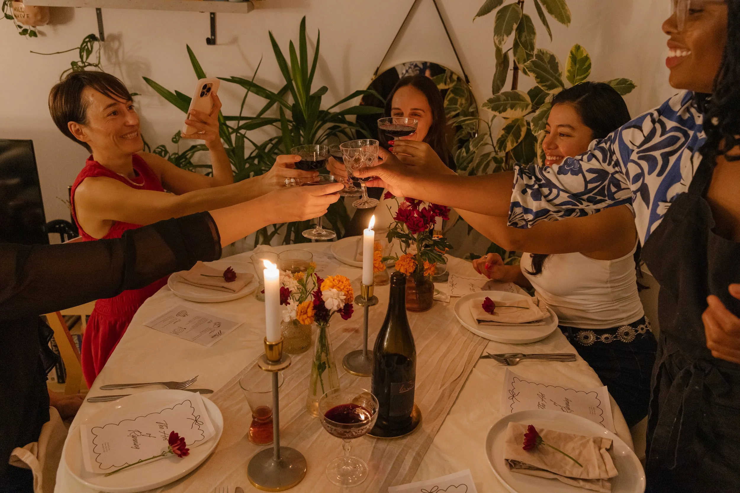 Five women are raising glasses in a toast at a dinner party, smiling and celebrating around a decorated table with candles and flowers at Dinner for One Supper Club..