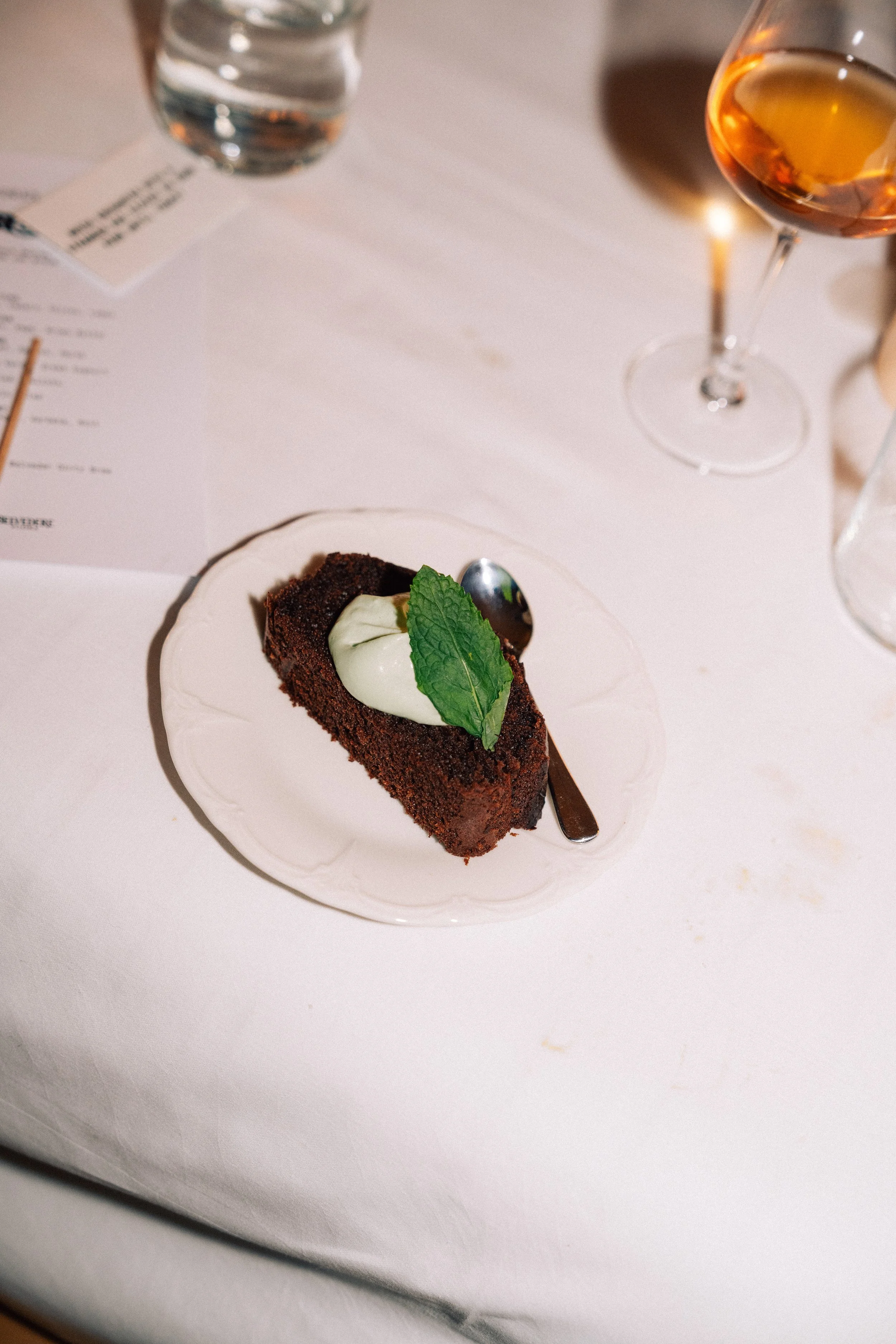 A slice of chocolate cake with a dollop of cream and a mint leaf on top, placed on a white plate, with a fork beside it on a white tablecloth. In the background, there are glasses of water and a glass of rosé wine, as well as a paper menu.