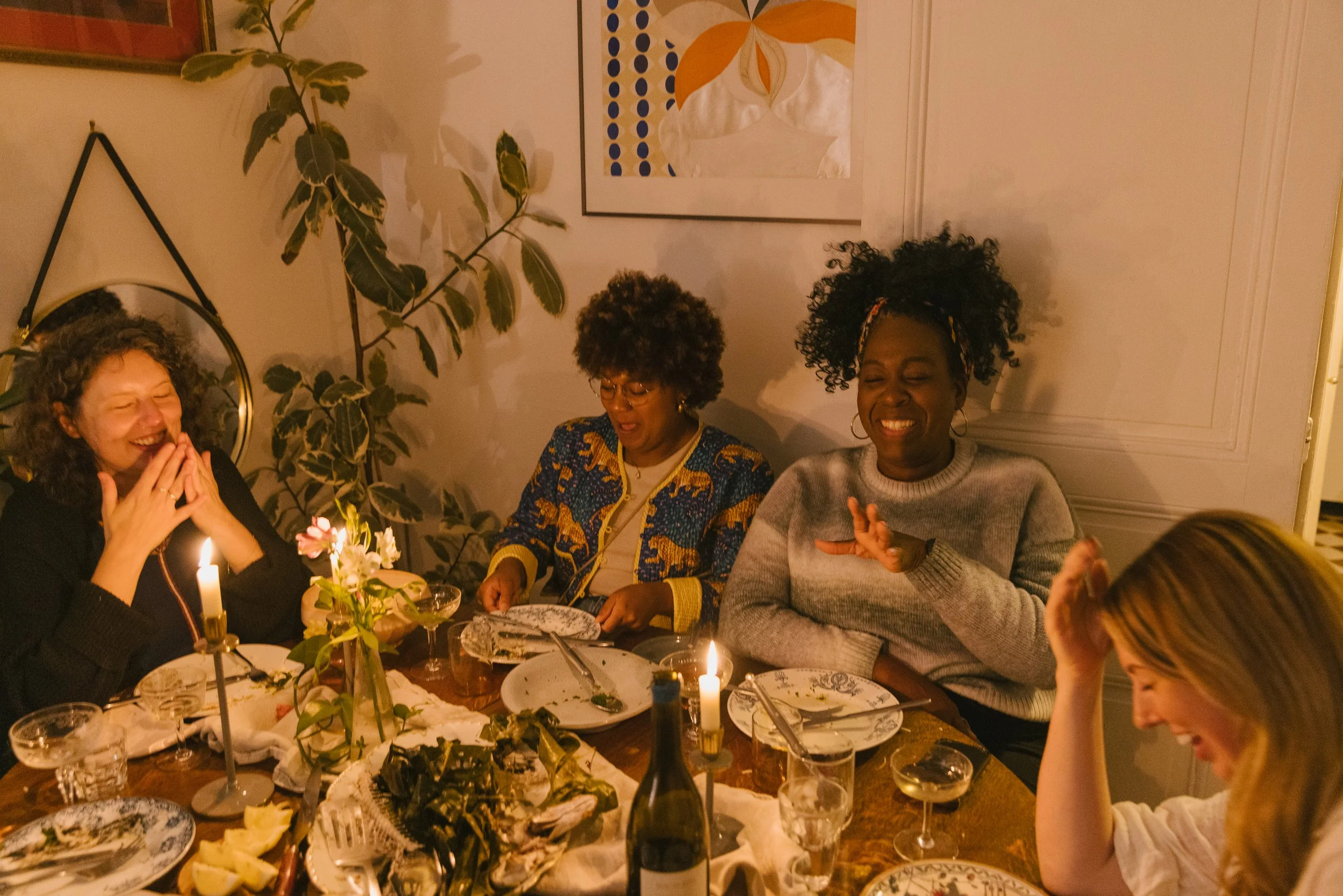 A group of four women enjoying a dinner for one supper clubat a table with candles, flowers, and food. They are smiling and laughing, surrounded by tableware and drinks, in a warmly lit room decorated with wall art and plants.