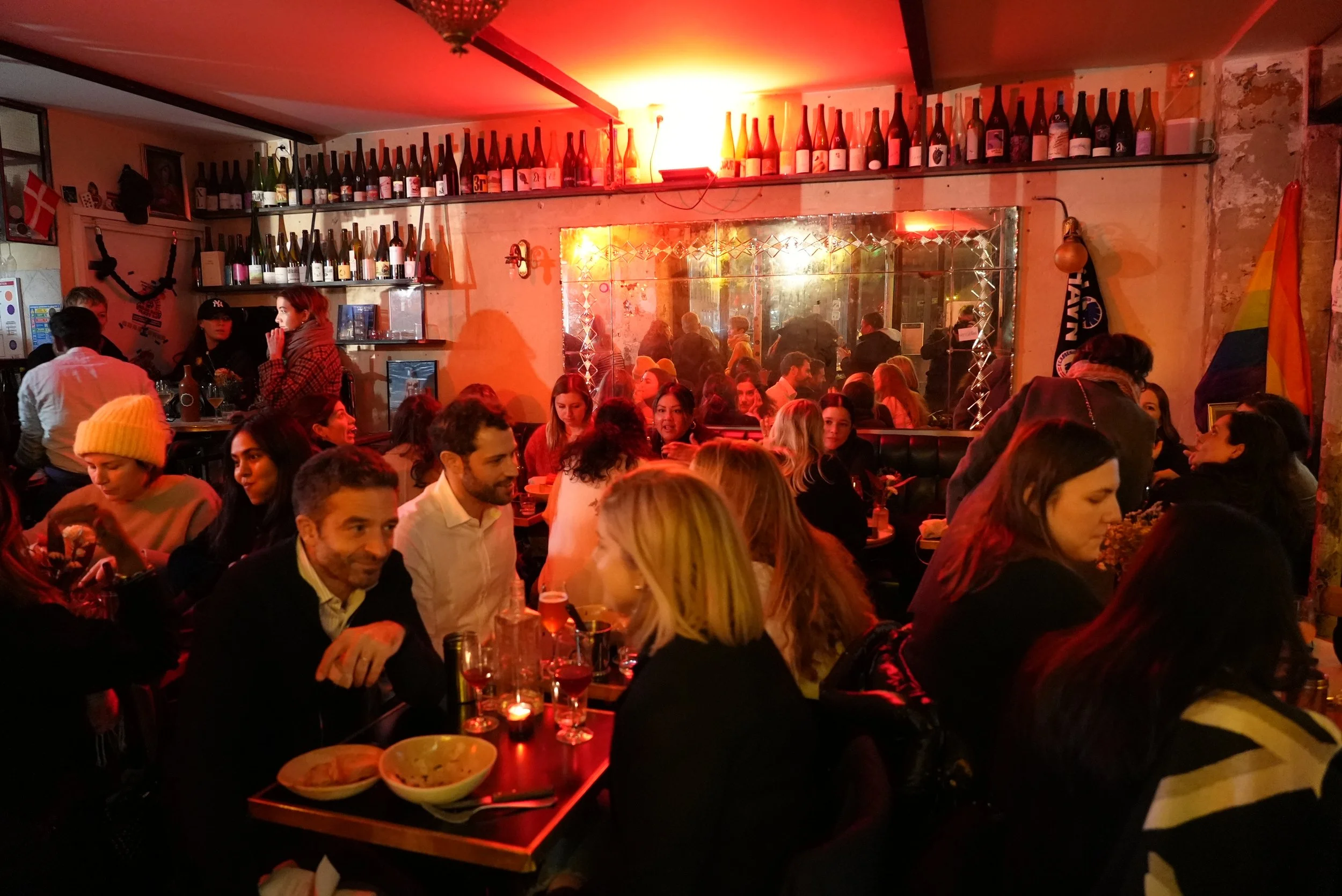 A lively bar or restaurant with many people sitting at tables, enjoying drinks and socializing. The room has warm red lighting, a mirror on the wall, and shelves with bottles of wine and champagne.