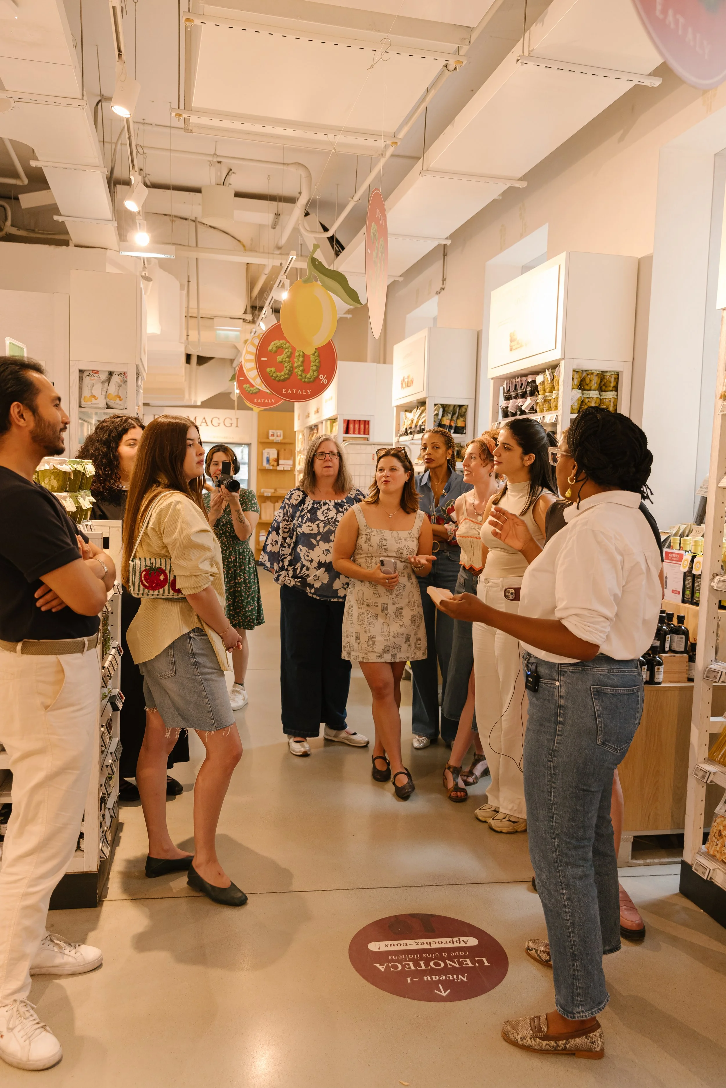 Group of people gathered inside a store, listening to a woman speaking, with shelves of products behind them and signs hanging from the ceiling.