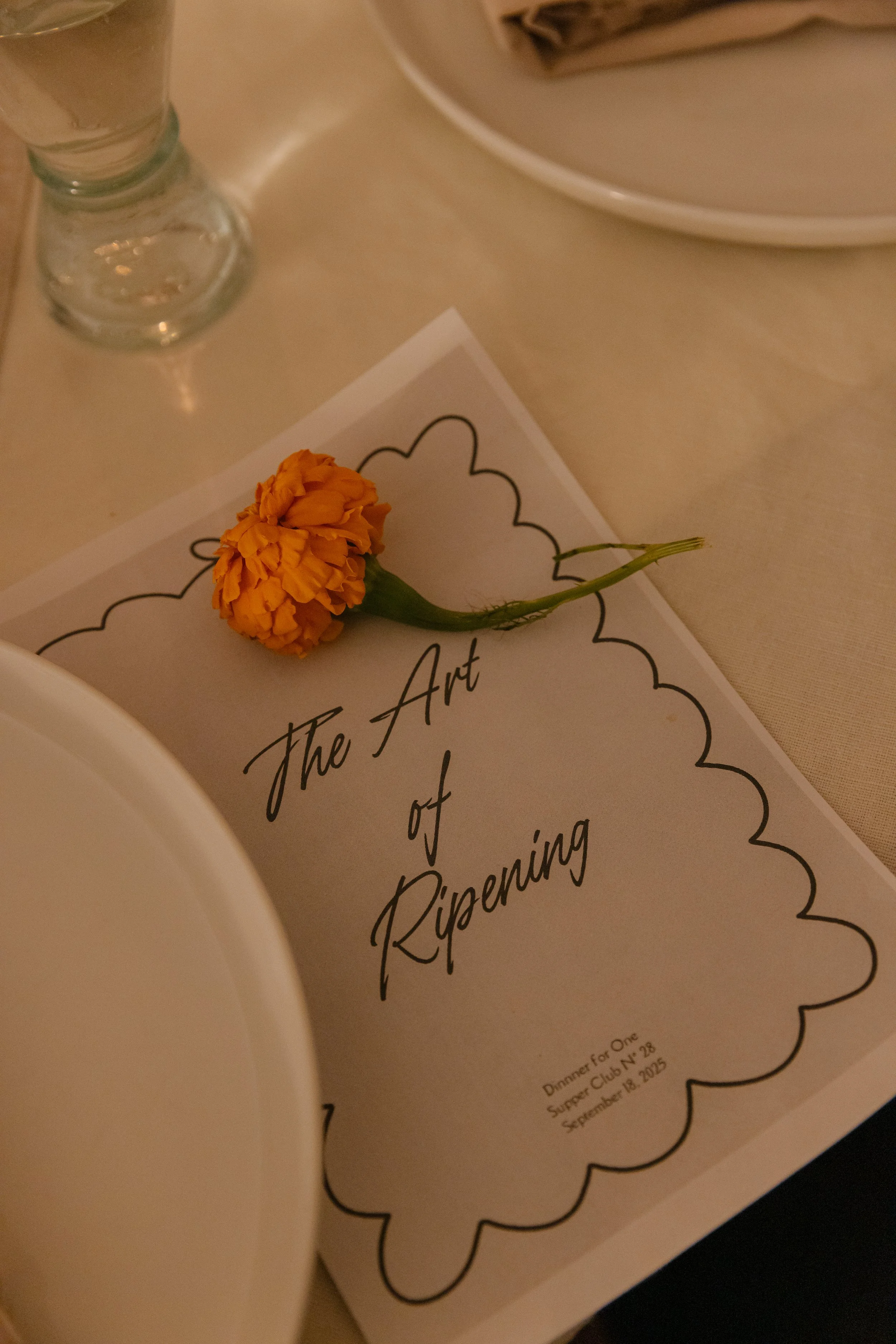 A menu titled 'The Art of Risenning' with a small orange flower resting on top, placed on a table with a partially visible glass and plate at Dinner for One Supper Club..