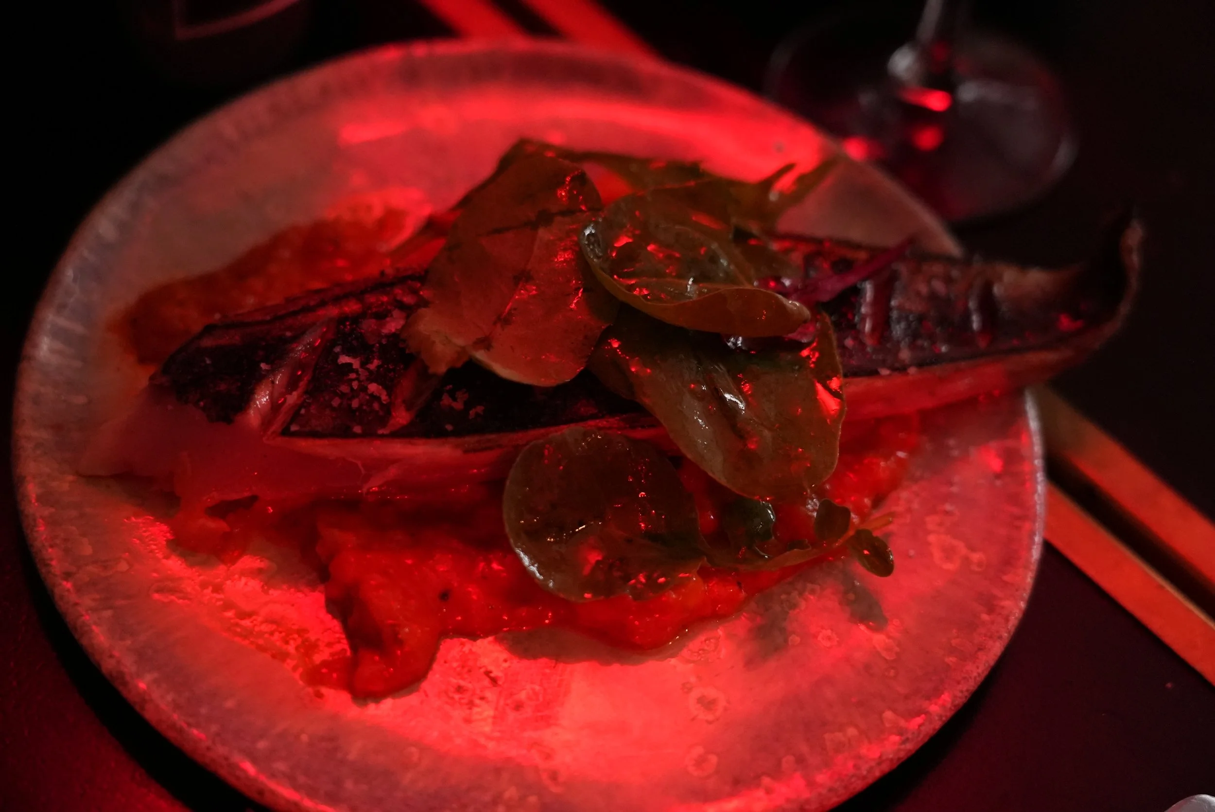 A plate with grilled fish topped with seaweed and garnished with thinly sliced vegetables or herbs, illuminated by moody red lighting in a dark setting.