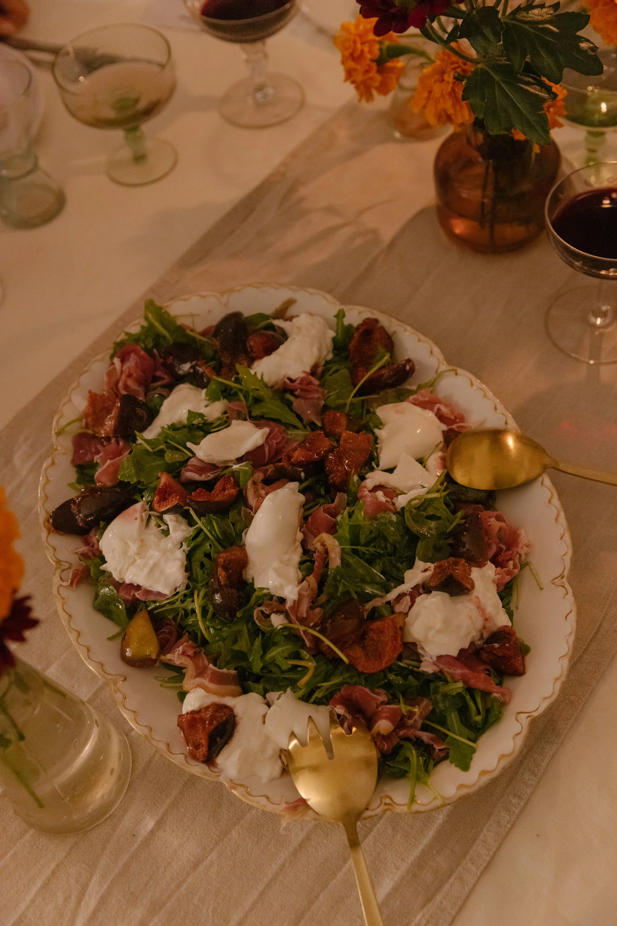 A large serving platter with a mixed green salad topped with mozzarella cheese, sun-dried tomatoes, prosciutto, and olives, with a gold-colored serving spoon. In the background, there are glasses of red and white wine, a floral centerpiece with orang