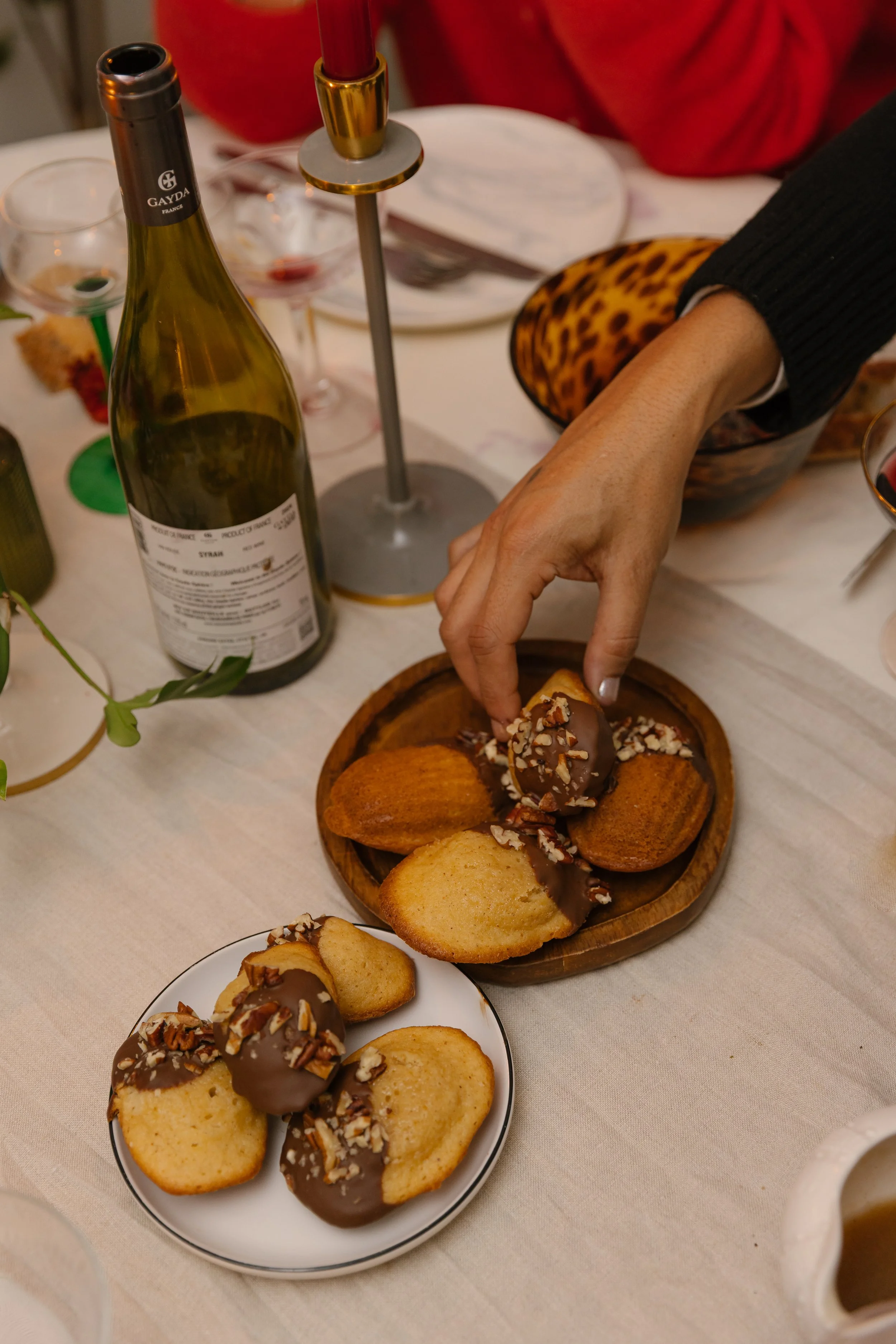 A table set with cookies topped with chocolate and nuts, a wine bottle, wine glasses, and a person reaching for a cookie.