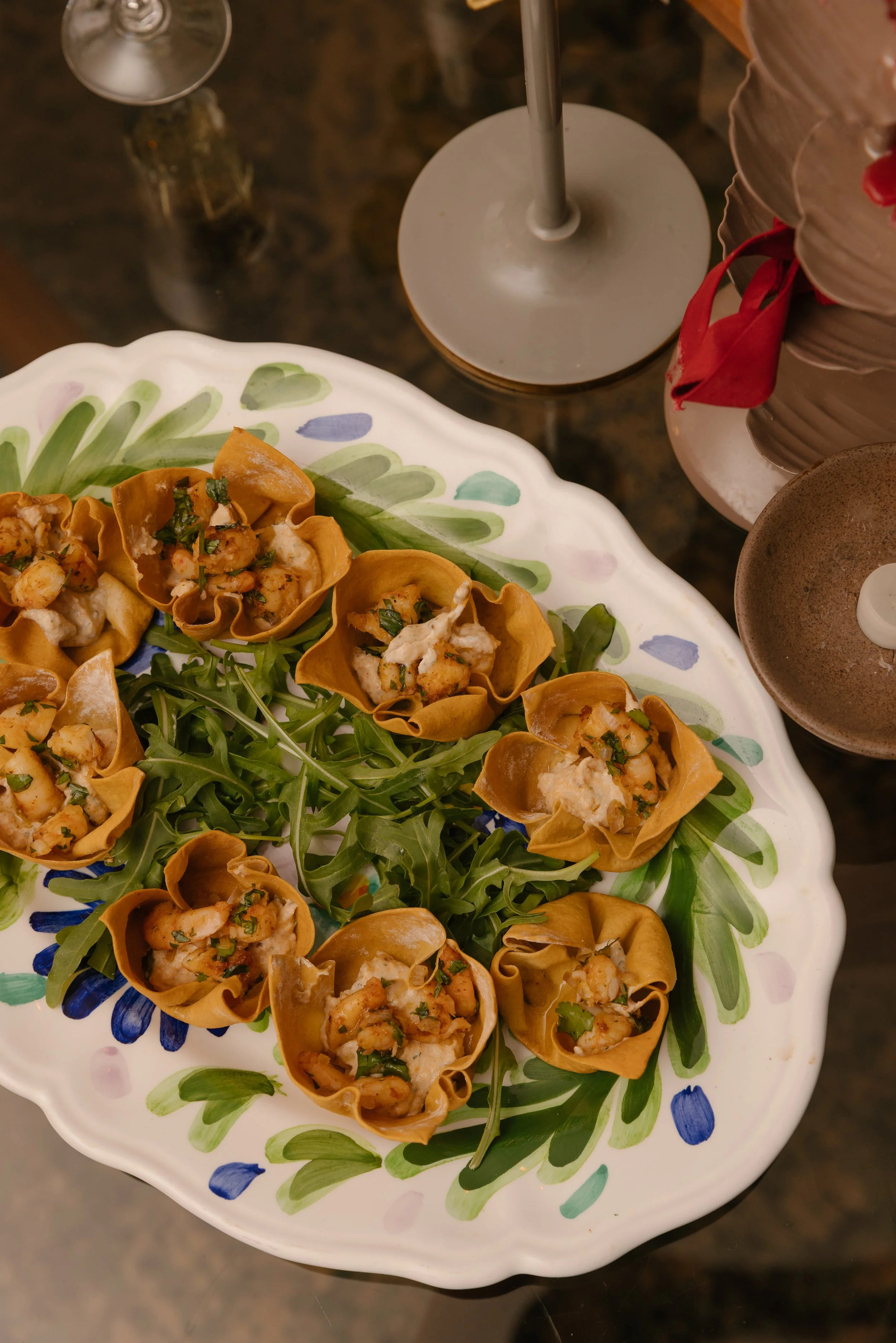 A decorative plate filled with small, golden-brown pasta cups topped with cooked shrimp, herbs, and a creamy sauce. The plate has a colorful, floral pattern with green, blue, and purple accents. In the background, there is a tall, slender silver cand