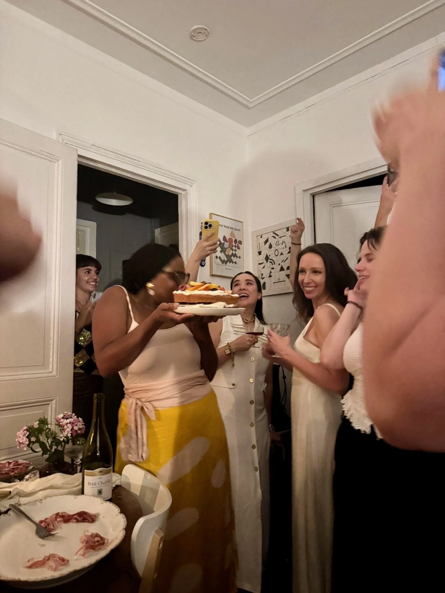 Group of friends celebrating around a table with food and cake in a cozy, white-walled room.
