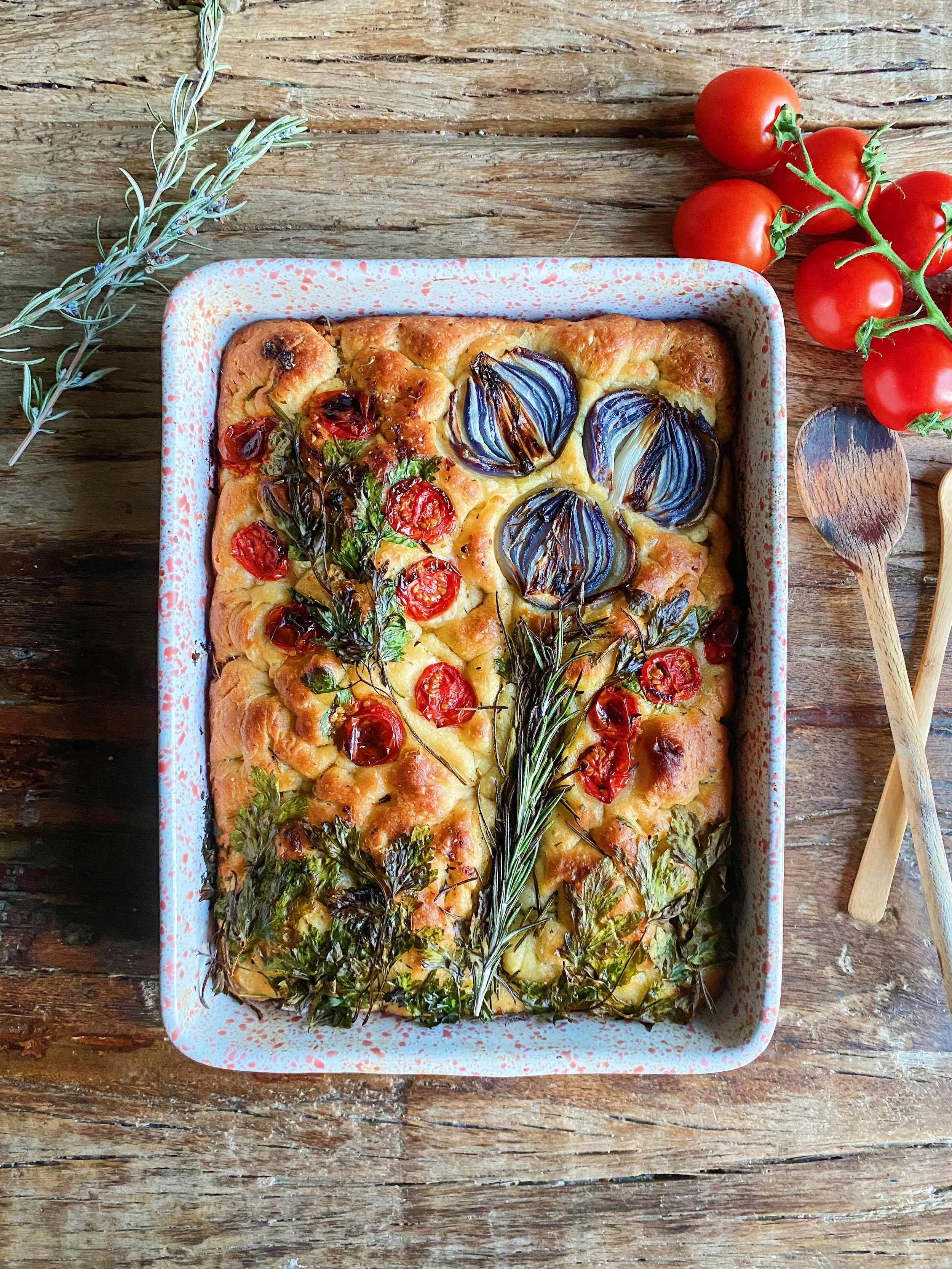 Homemade focaccia bread topped with roasted garlic, cherry tomatoes, and fresh herbs on a ceramic baking dish, placed on a rustic wooden table with a sprig of rosemary and cherry tomatoes nearby.