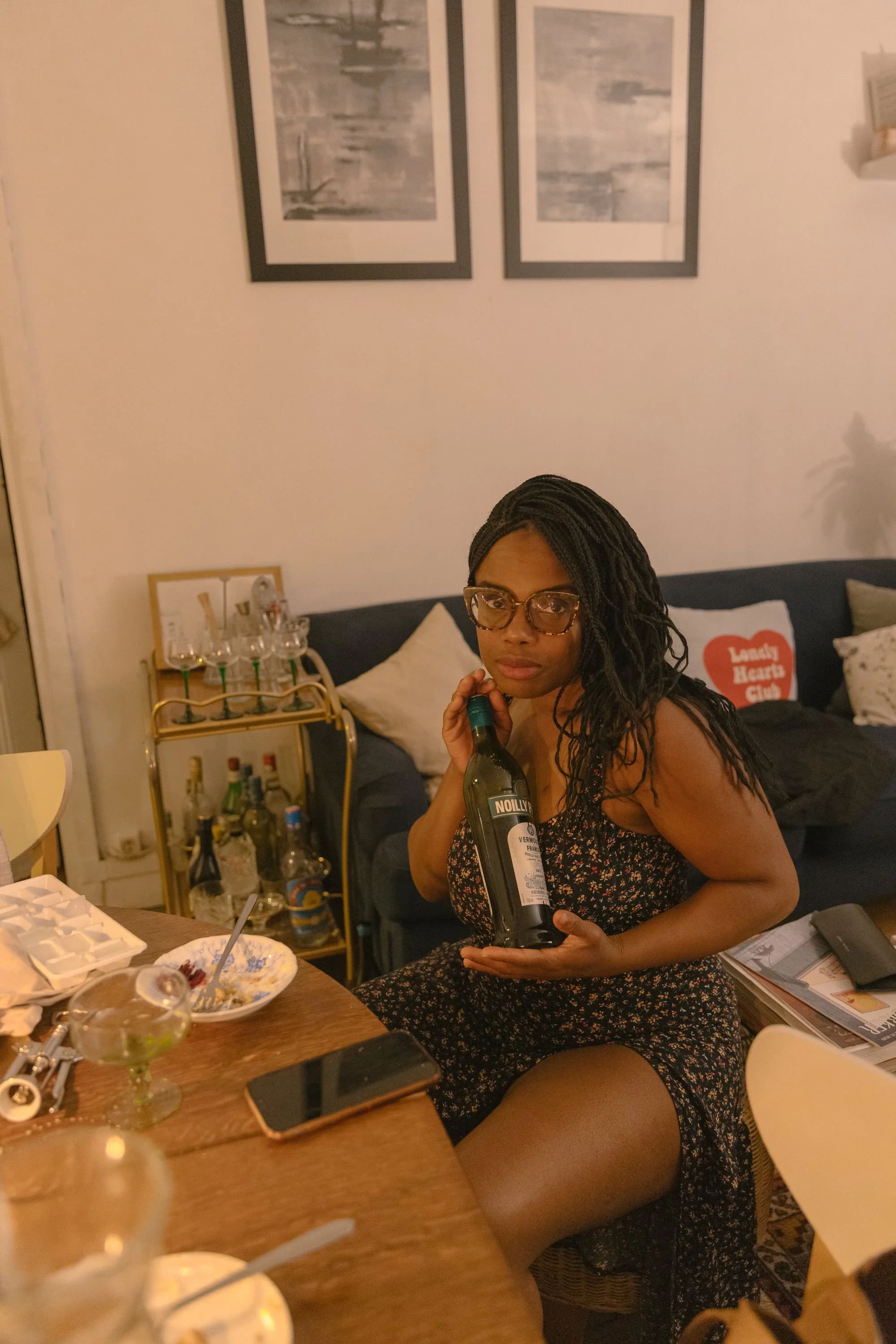A woman with long black twisted hair and glasses sitting on a couch, holding a bottle of wine in a cozy living room with decorative paintings and a bar cart background.