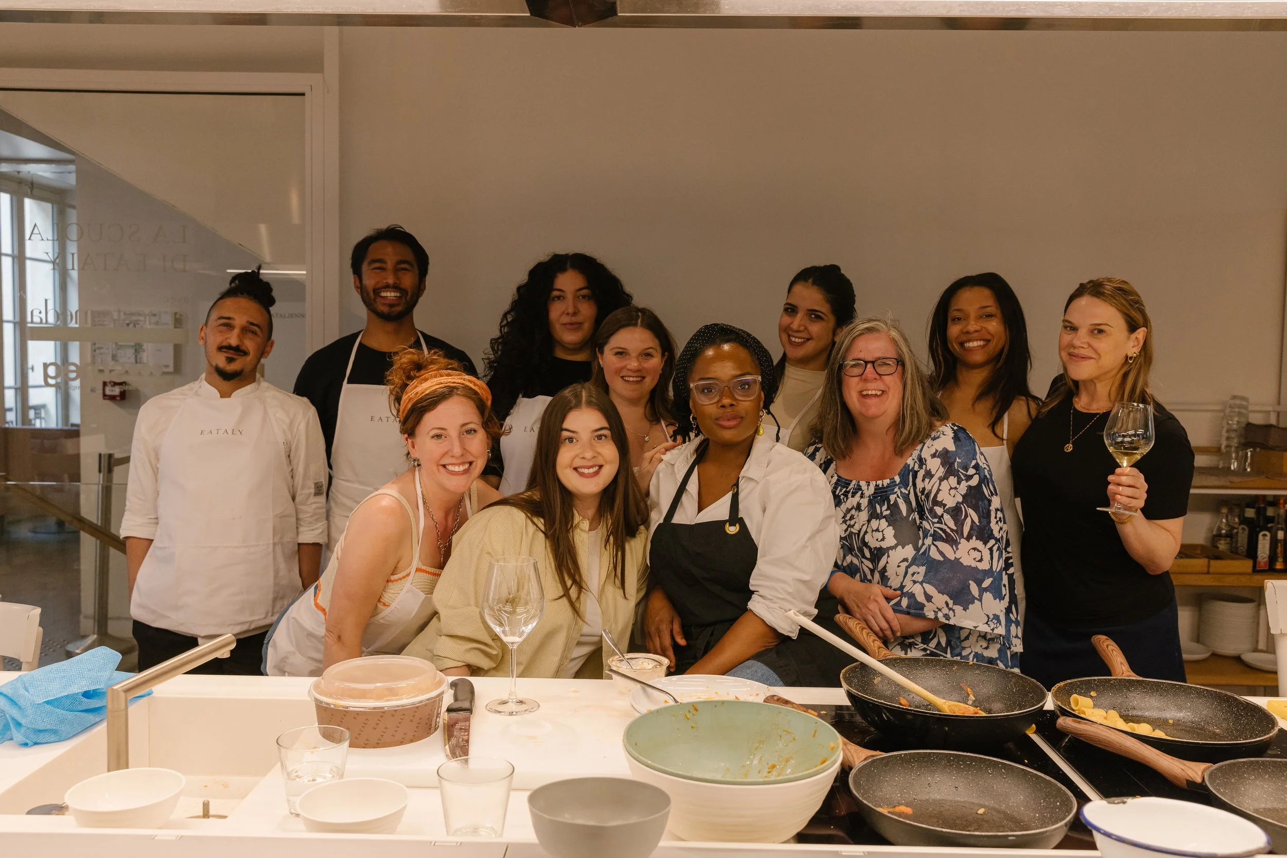 A diverse group of people in a restaurant kitchen, smiling and posing together after cooking a meal, with empty plates and cooking utensils on the counter in front of them.