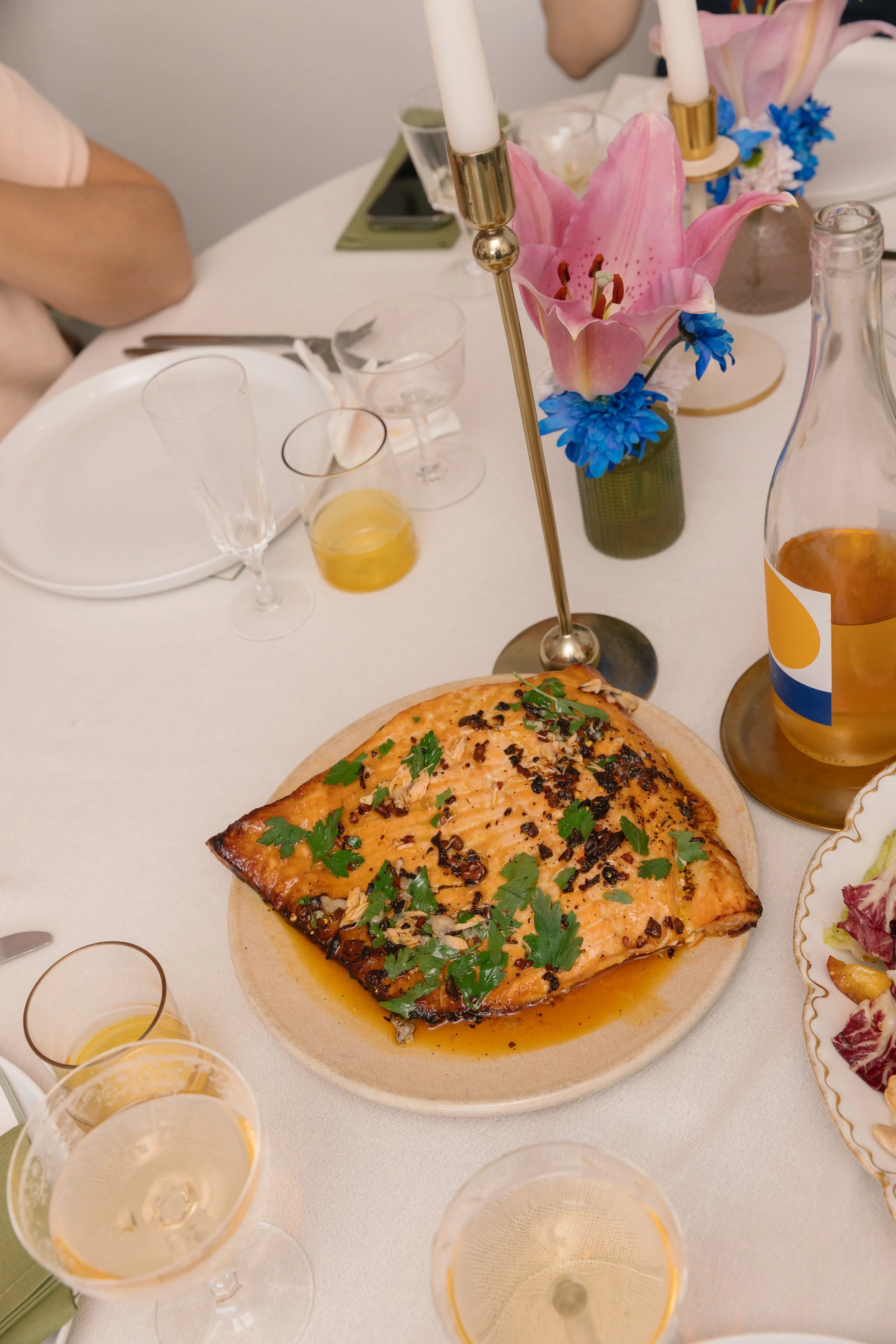 A table set for a meal with a focus on a cooked salmon fillet garnished with herbs, surrounded by various glasses and a vase with pink lilies and blue flowers.