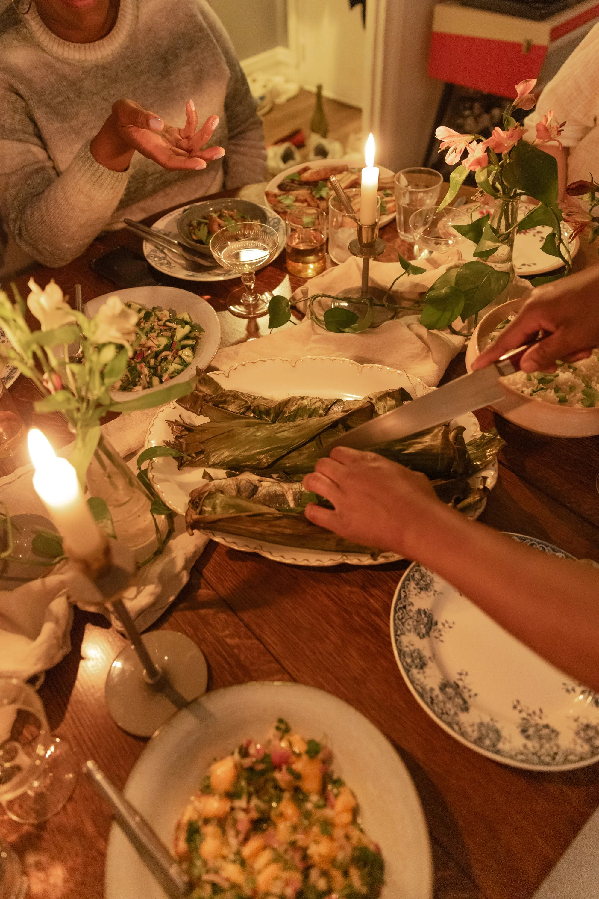 A dinner table set for a festive meal with various dishes, a lit candle, flowers, and people serving food in a cozy indoor setting.