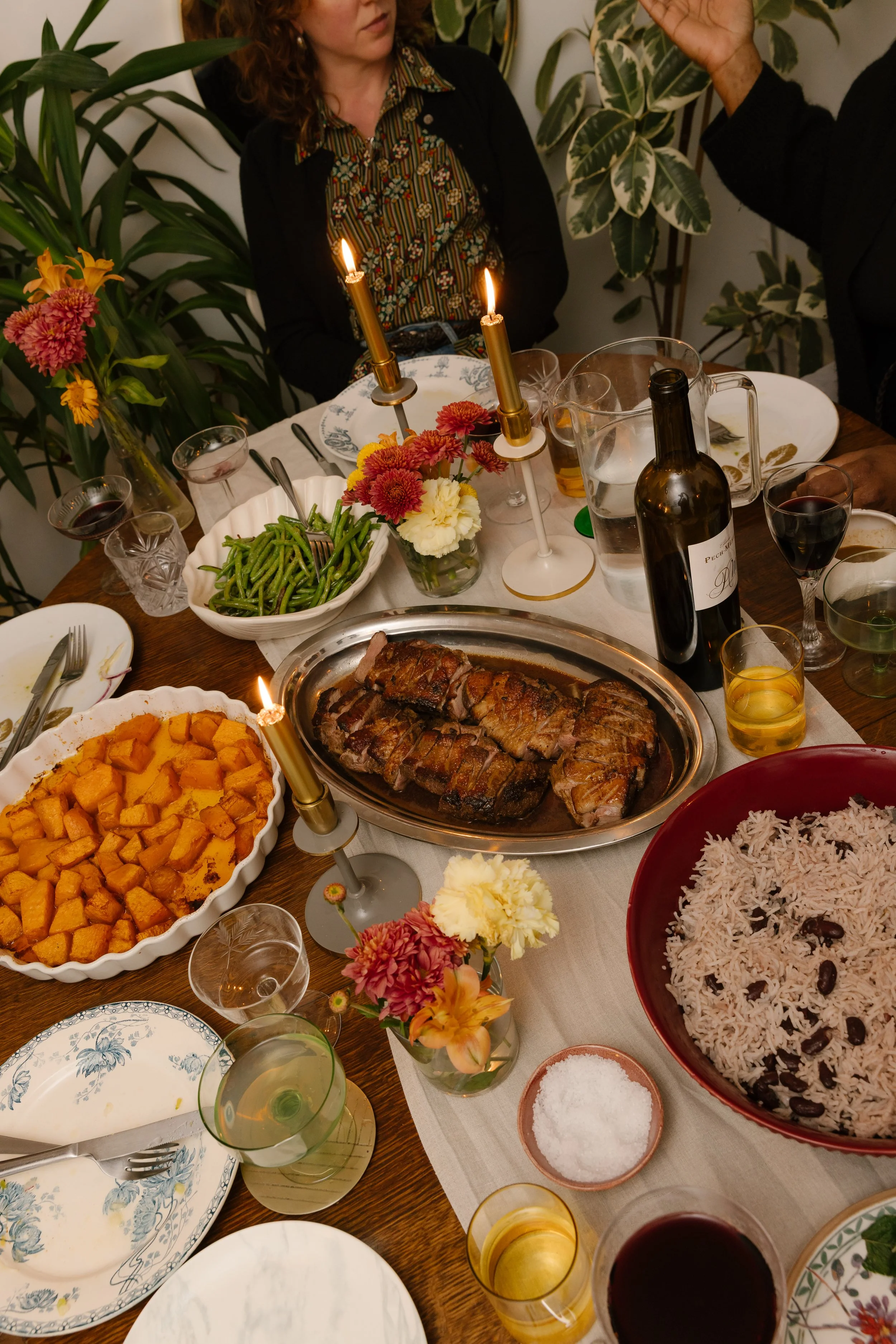 A dinner table with various dishes including a large platter of sliced grilled meat, bowls of rice and sweet potatoes, a green bean casserole, and a colorful floral centerpiece. There are also glasses of wine, candles, and decorative plates.