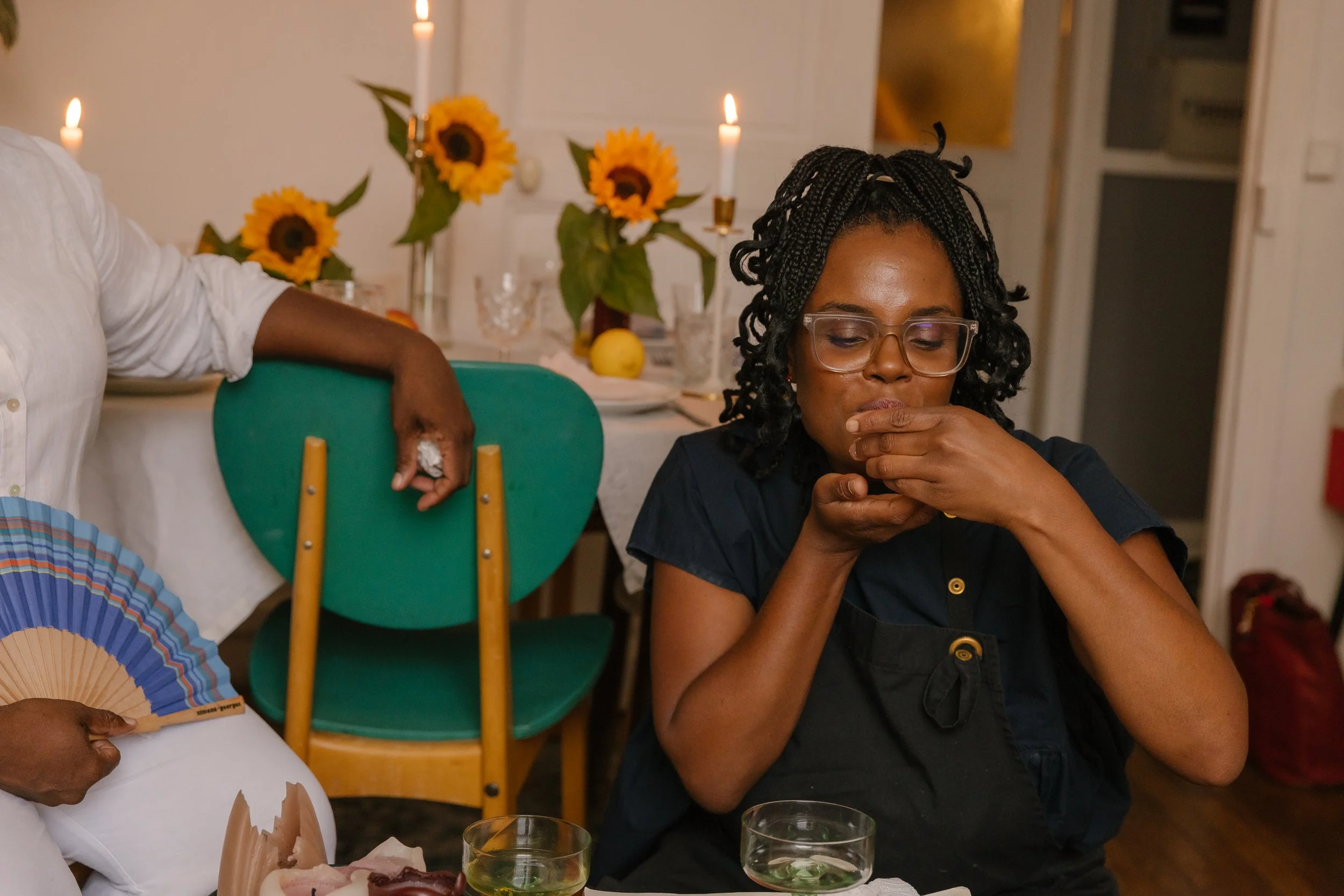 A woman with glasses and braids covering her mouth with her hands at a dinner table decorated with sunflowers, white candles, and various drinks.