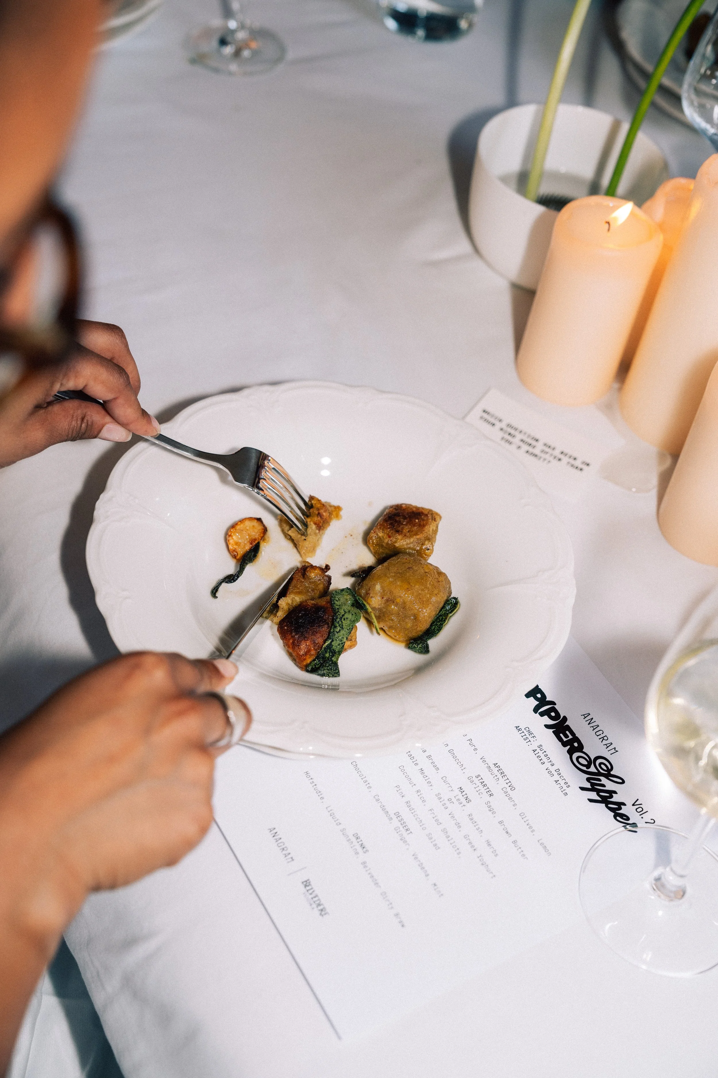 A person is cutting into food on a white decorative plate at a table with a menu, candles, and wine glasses.