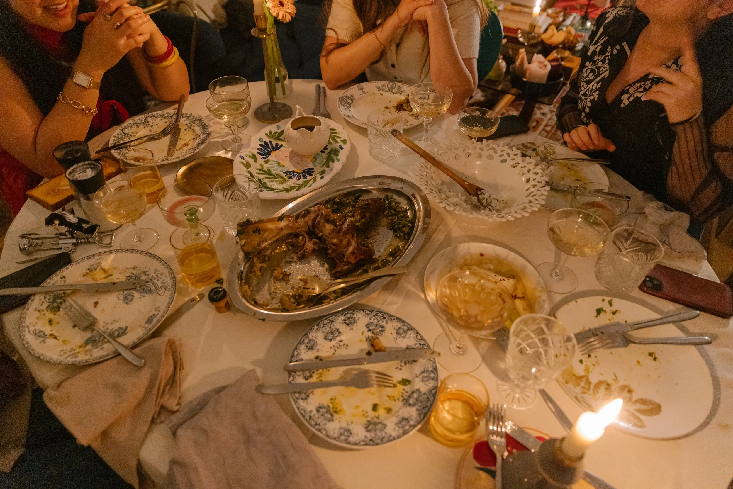 A dining table after a meal with several empty and partially eaten plates, glasses of wine and drink, utensils, a white doily, a flower vase, and a candle, surrounded by people who are socializing.