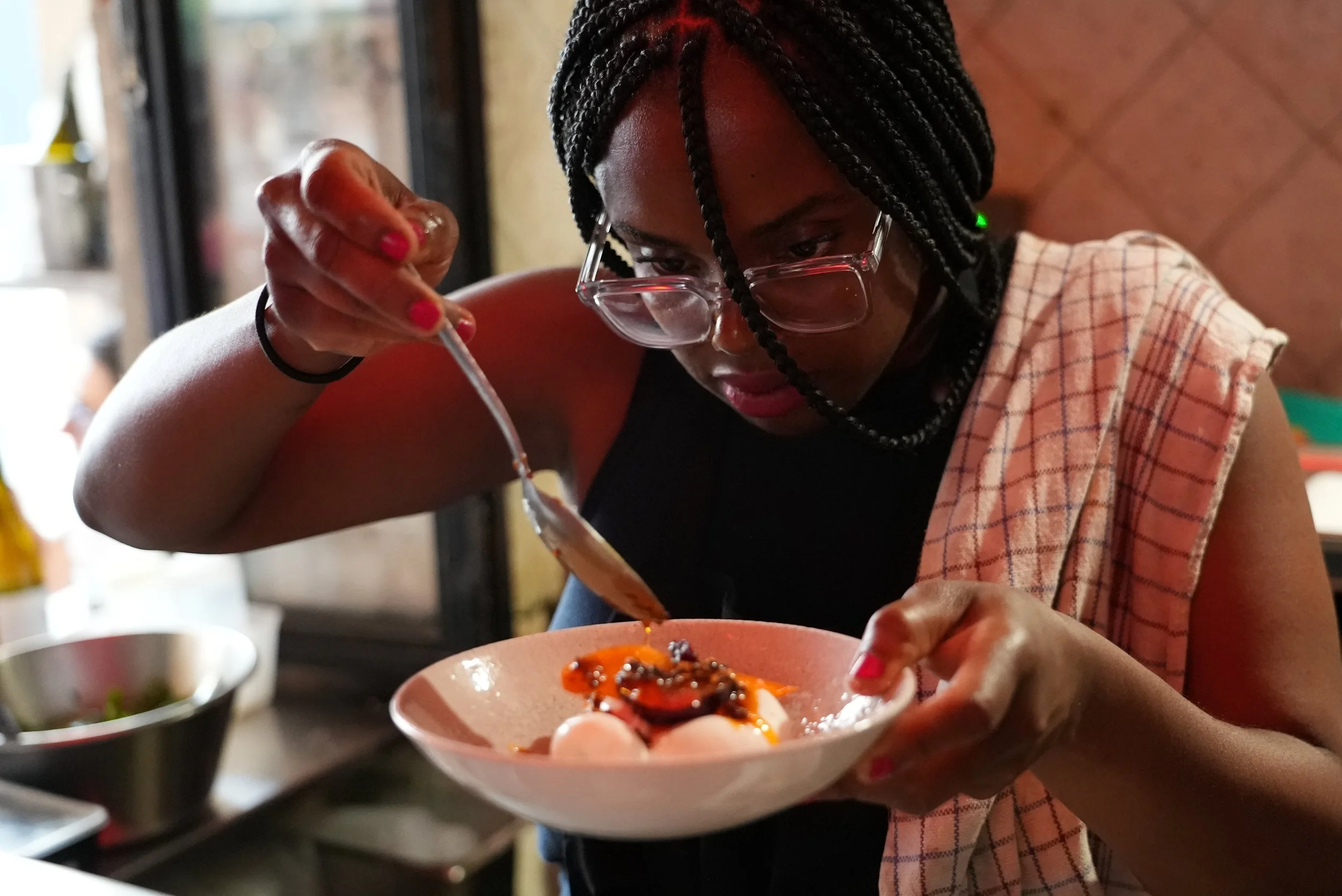 Sutanya Dacres with braided hair, glasses, and a towel over her shoulder is carefully spooning sauce onto a bowl of food in a restaurant.