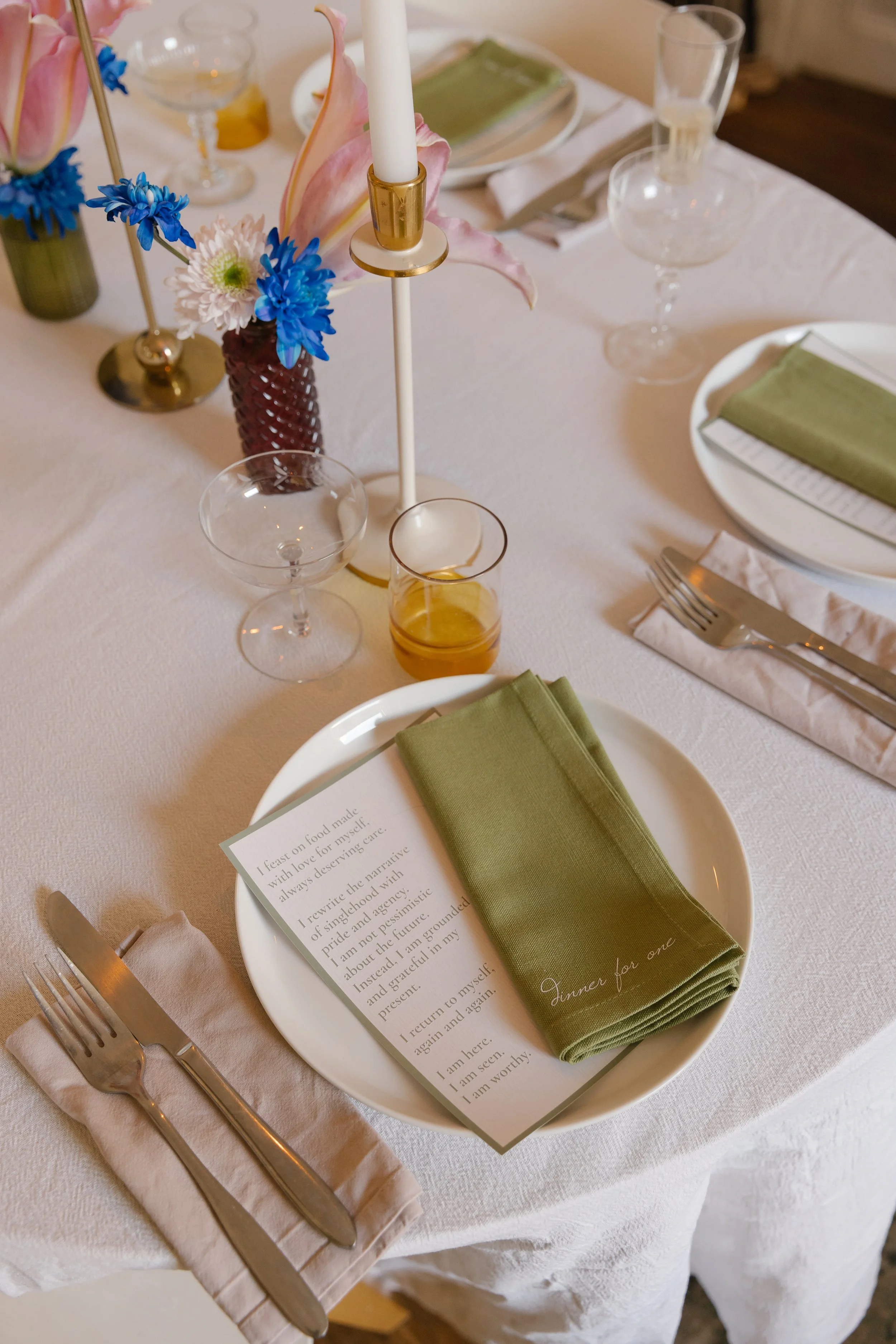 A formal table setting with a white tablecloth, green napkins, and clear glassware. A centerpiece with pink and white flowers, including blue and white blooms, is on the table. Silverware is arranged on a napkin, and a white plate holds a green napki