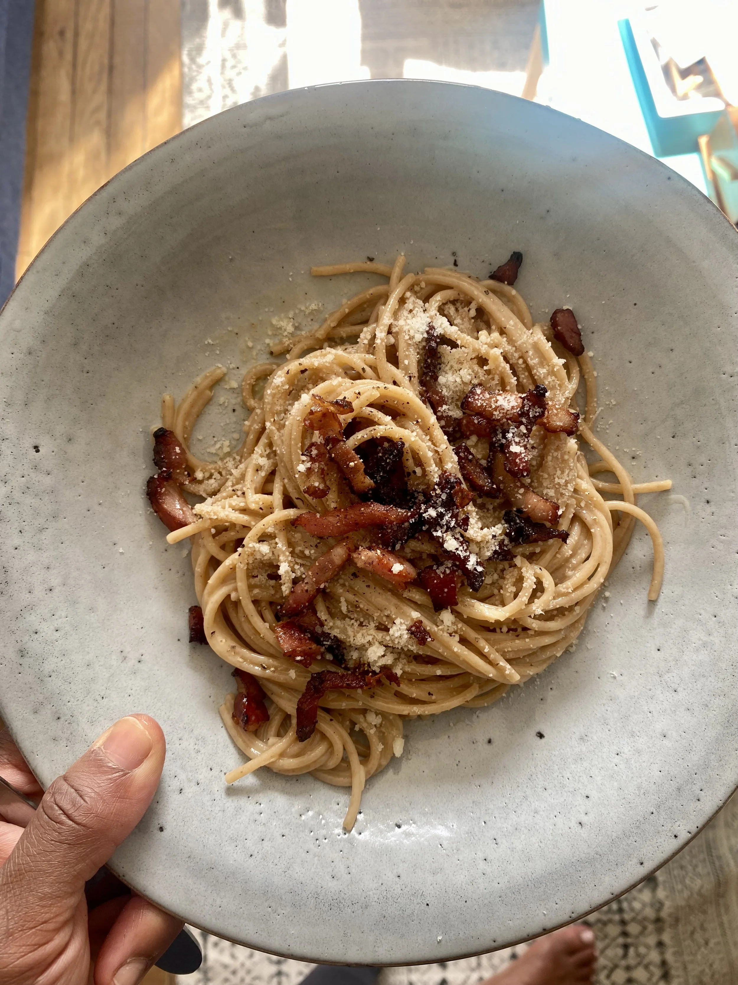 A plate of spaghetti with bacon bits and grated cheese, held by a person's hand with a thumb visible.