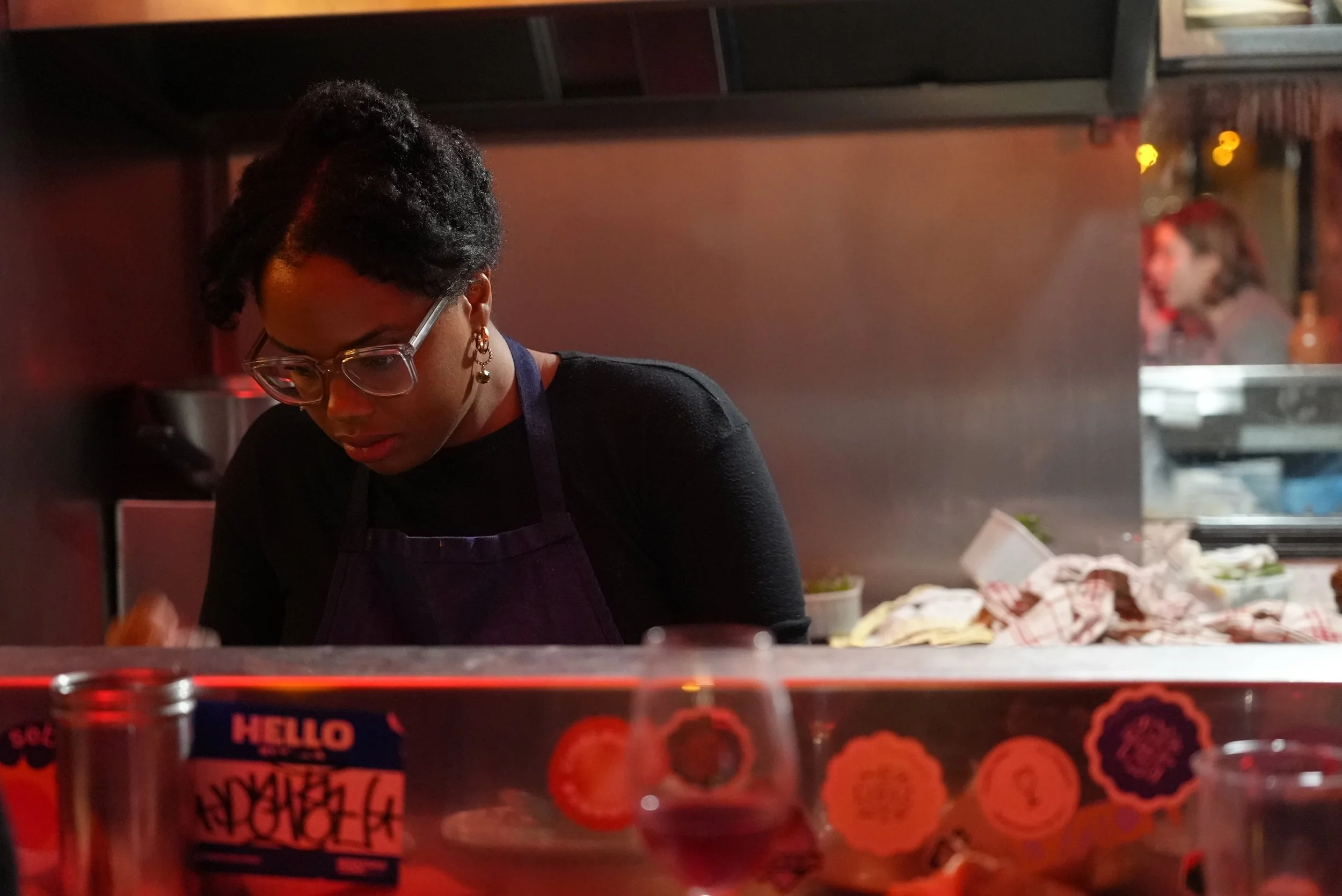 Sutanya Dacres with short curly hair, wearing glasses, earrings, a black shirt, and a dark apron, preparing food in a restaurant kitchen with warm lighting.