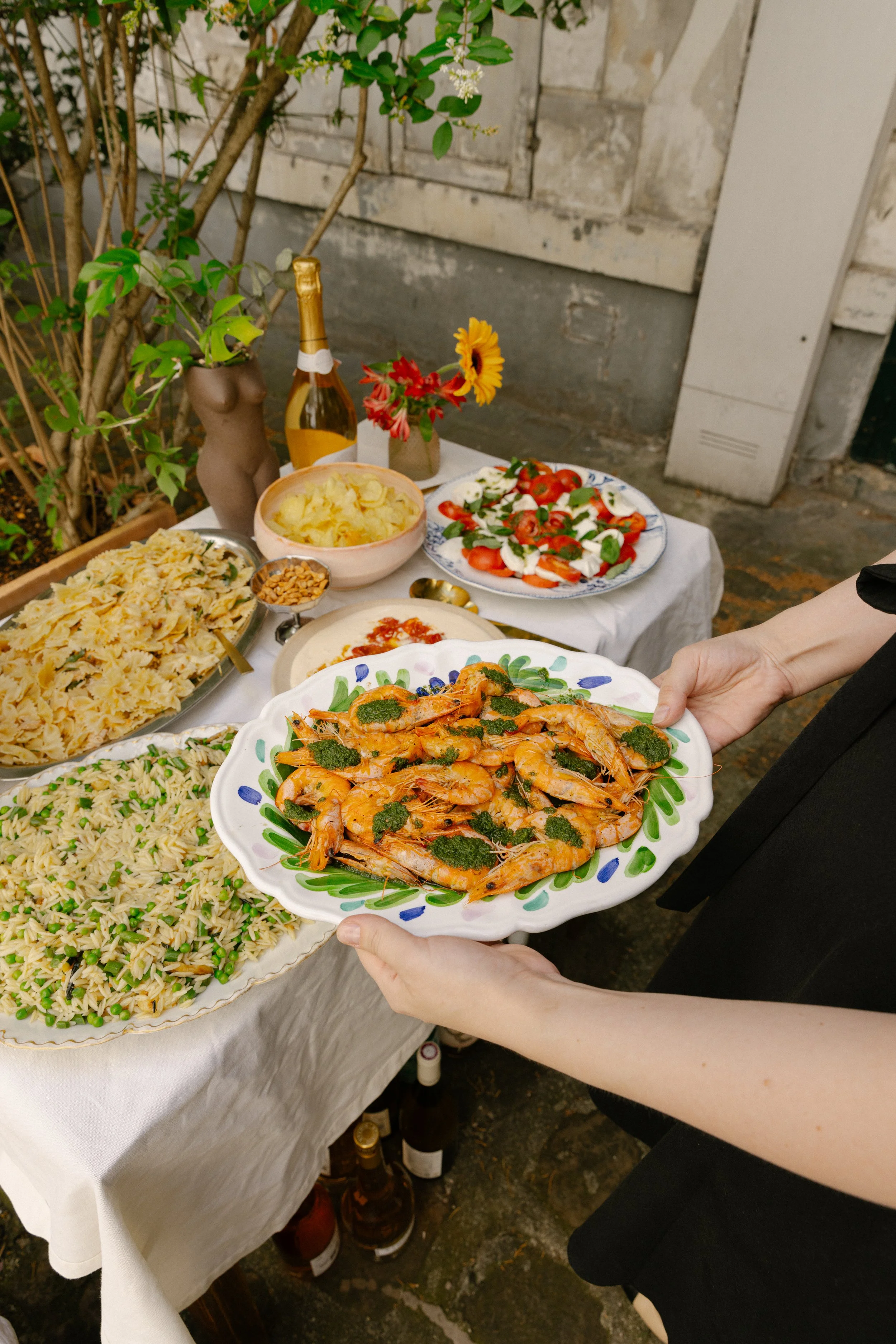 A person holding a white plate of cooked shrimp with herbs at an outdoor gathering table with various dishes, a bottle of champagne, and a vase with yellow and red flowers, set against a stone wall and greenery.