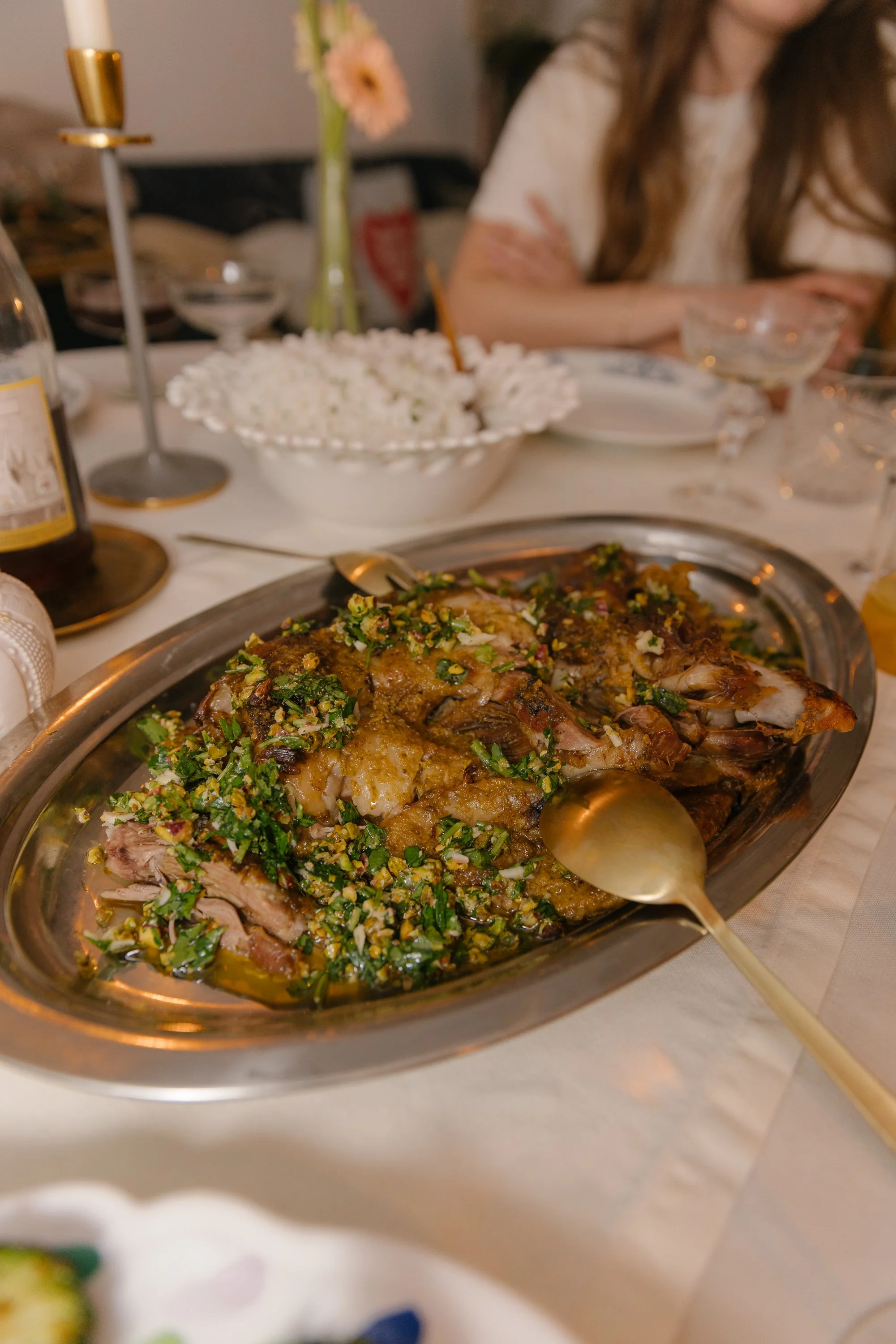A plated cooked fish garnished with herbs and chopped nuts, on a silver serving dish, with a golden spoon resting on the side of the dish. In the background, a table setting with various dishes, a bowl of rice, flowers, and a person with long brown h