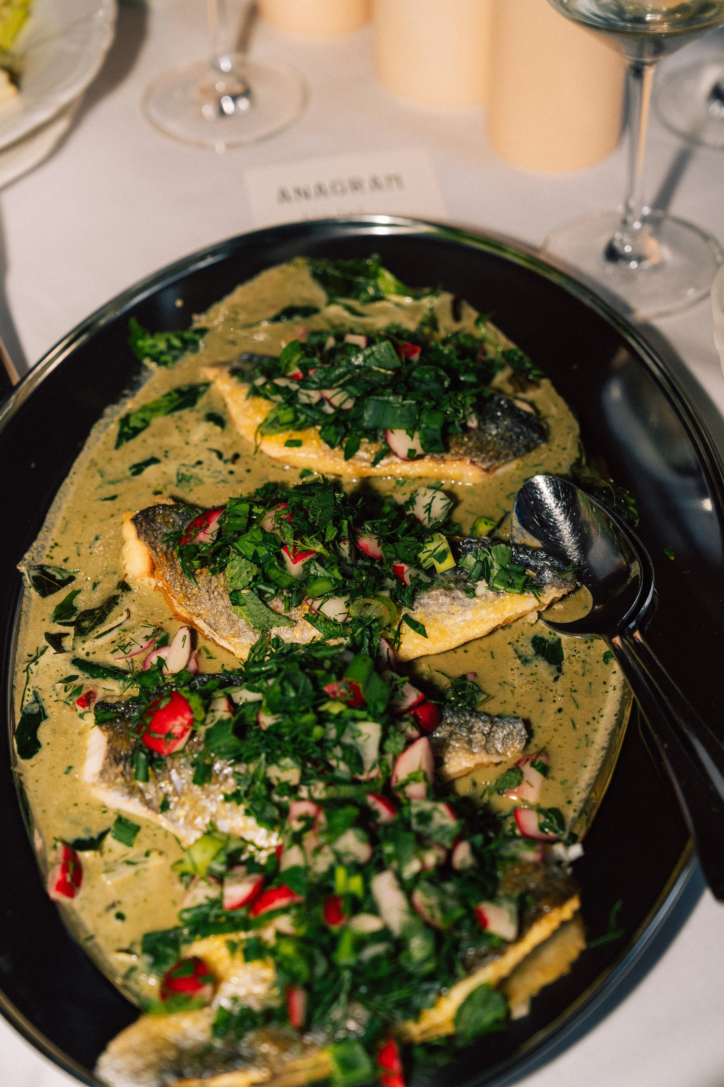 A fish dish with green herbs, radishes, and a creamy sauce in a black serving dish, with glasses and candles on a white tablecloth in the background.