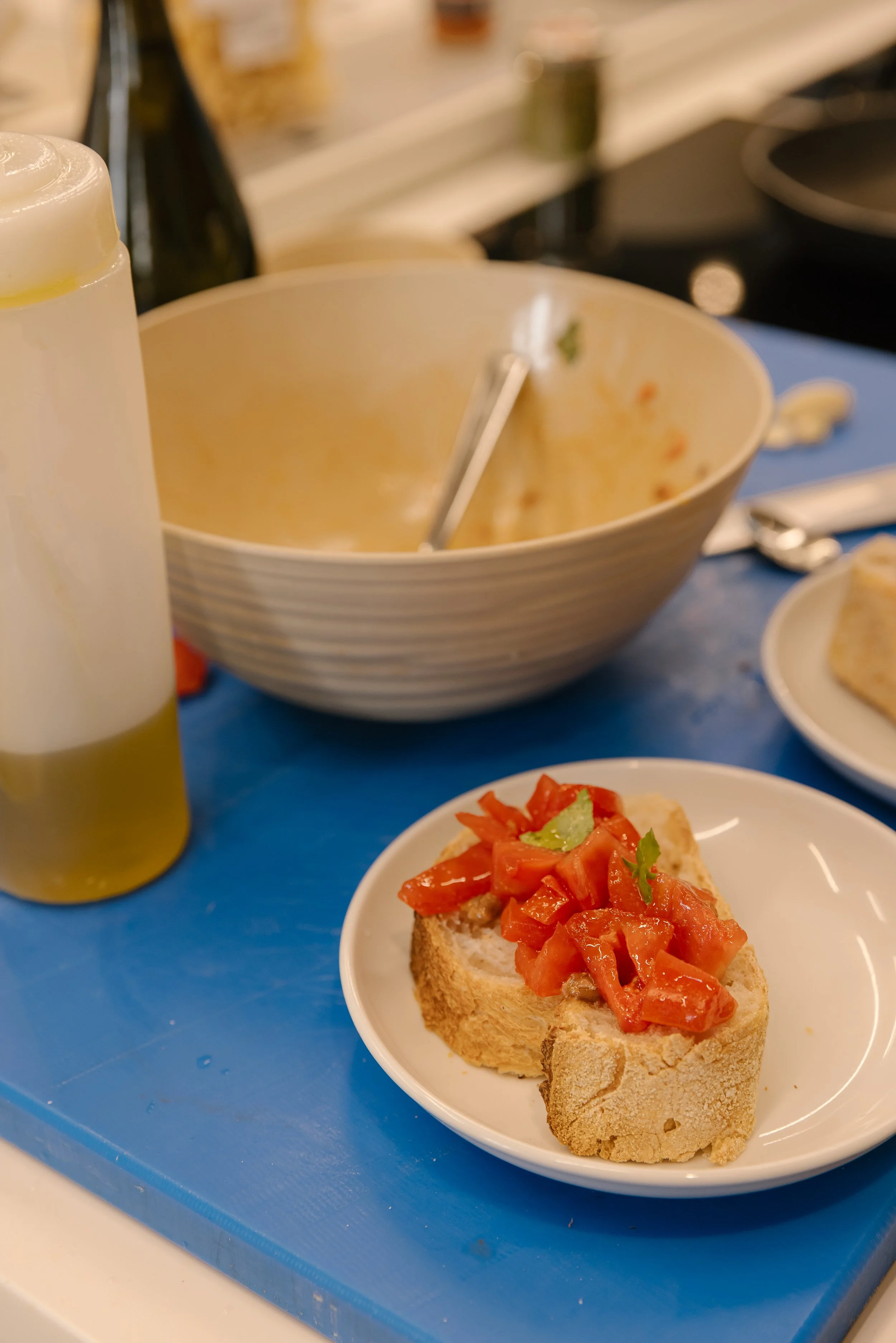 A plate with toasted bread topped with chopped tomatoes and herbs on a blue table. In the background, there is an empty bowl, a bottle, and part of a kitchen setting.