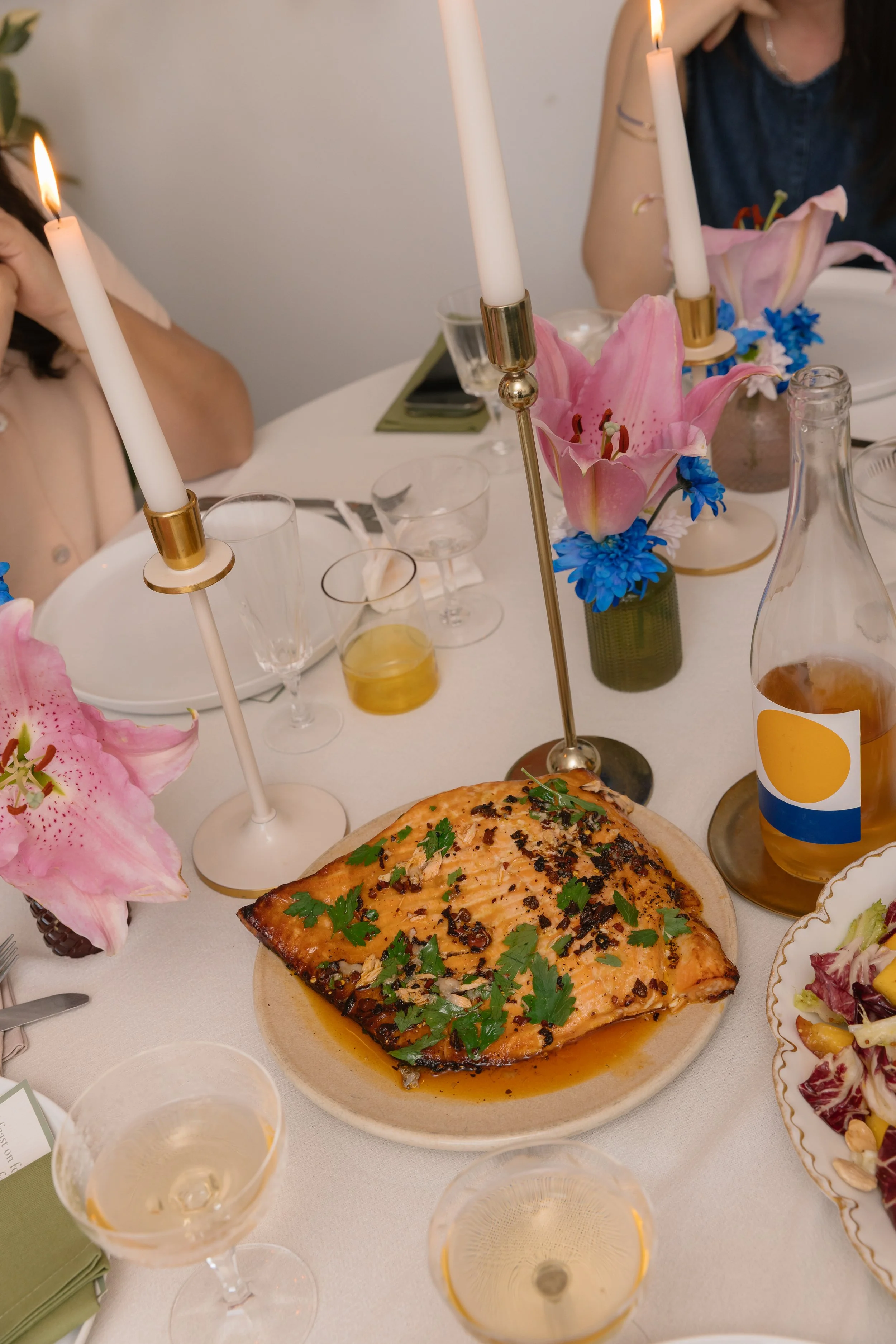 A dinner table with a plate of baked salmon garnished with herbs, surrounded by wine glasses, floral centerpieces with pink lilies and blue flowers, candlesticks with lit white candles, bottles, and various dishes.