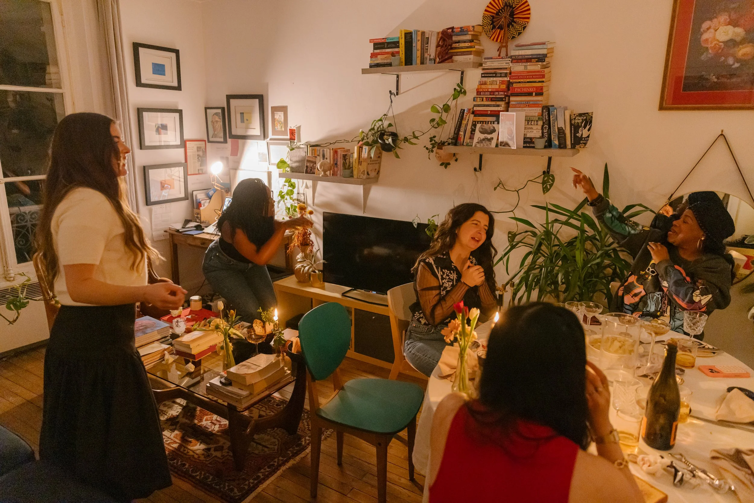 A group of women gathered in a cozy, warmly lit living room, engaged in conversation and celebration, with a table set with food and drinks, featuring books, artwork on the walls, and abundant indoor plants.