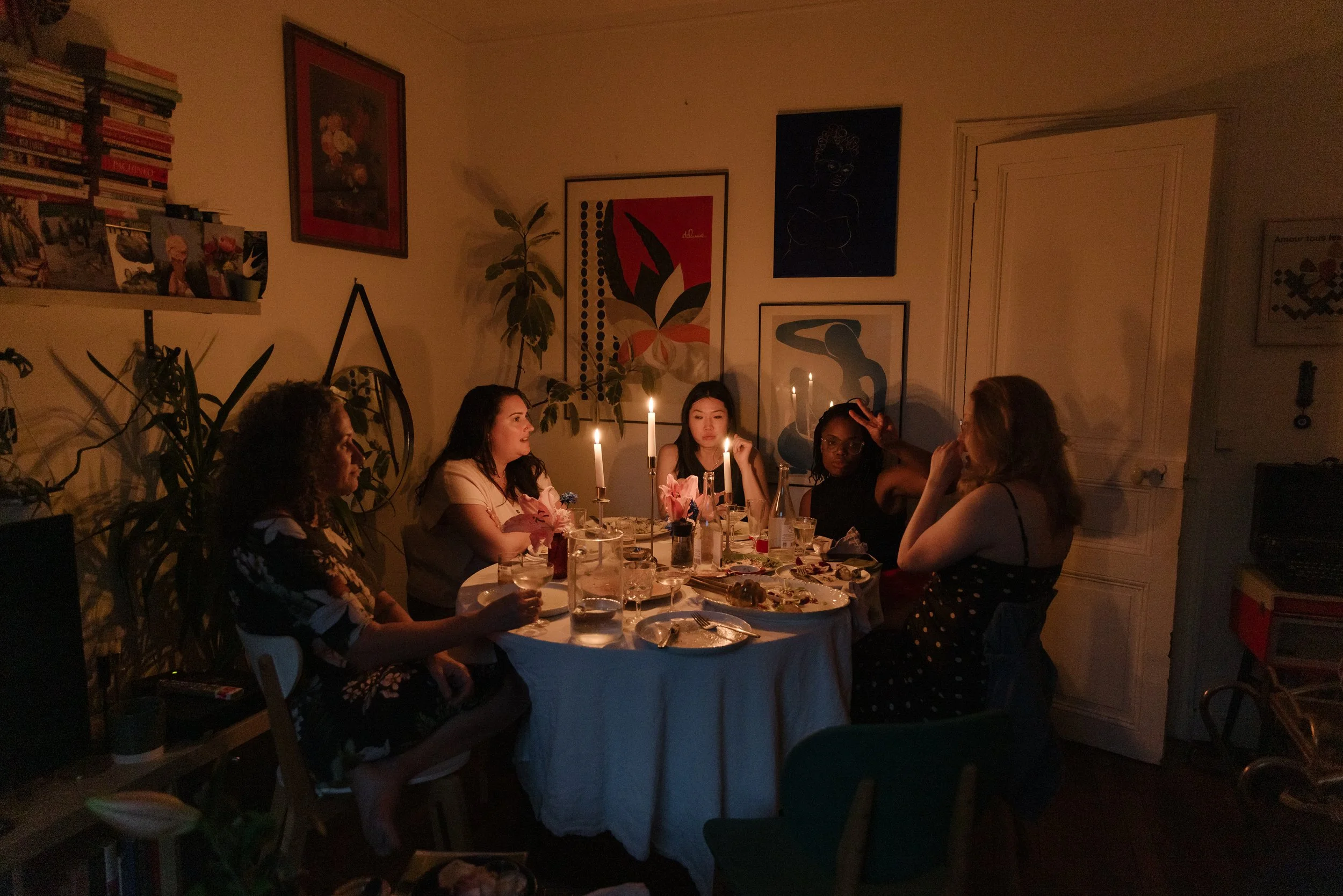 A group of women sitting around a dinner table in a dimly lit room decorated with framed artwork and potted plants, enjoying a meal.