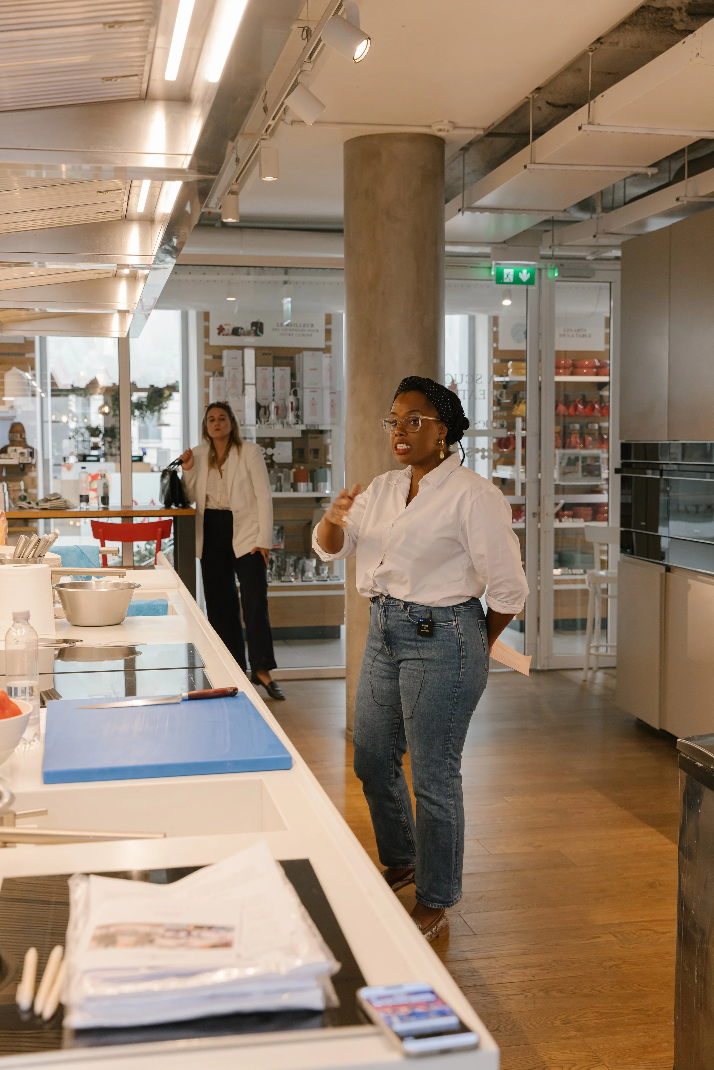 A woman wearing glasses and a white shirt is speaking or explaining, standing in a modern kitchen showroom. Another woman in a white blazer is in the background walking with a black bag. The space includes a large concrete pillar, ceiling lights, and