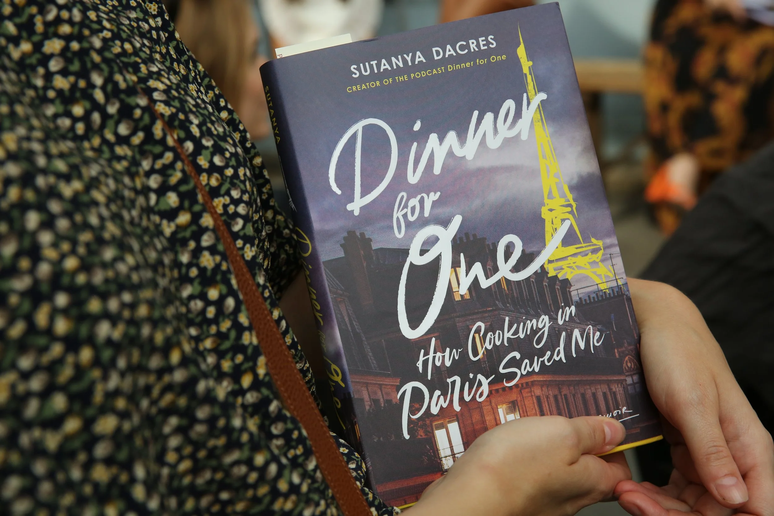 Close-up of a person holding a book titled "Dinner for One" by Sutanya Dacres. The person is wearing a floral patterned shirt and has a brown bag strap over their shoulder. In the background, there are other people and a blurred indoor setting.