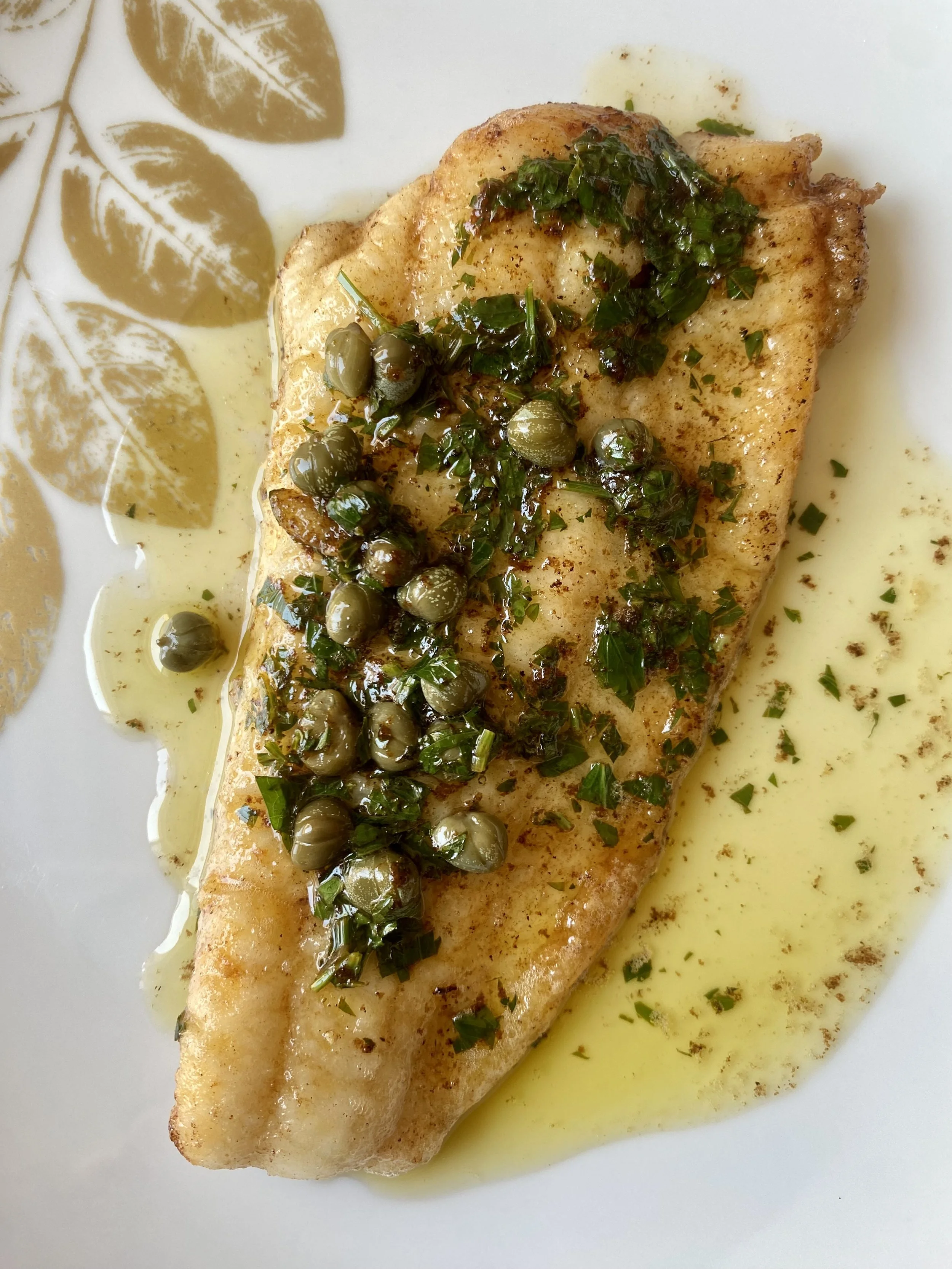 Pan-seared fish fillet topped with chopped herbs and capers, drizzled with lemon butter sauce, served on a white plate with a decorative gold leaf pattern.