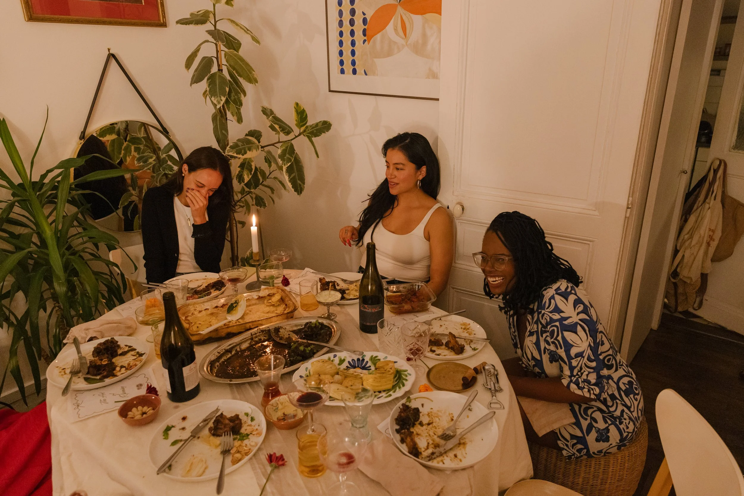 Three women at Dinner for One Supper club laughing and smiling. The table is filled with partially eaten food, drinks, and a lit candle. The women appear to be enjoying themselves