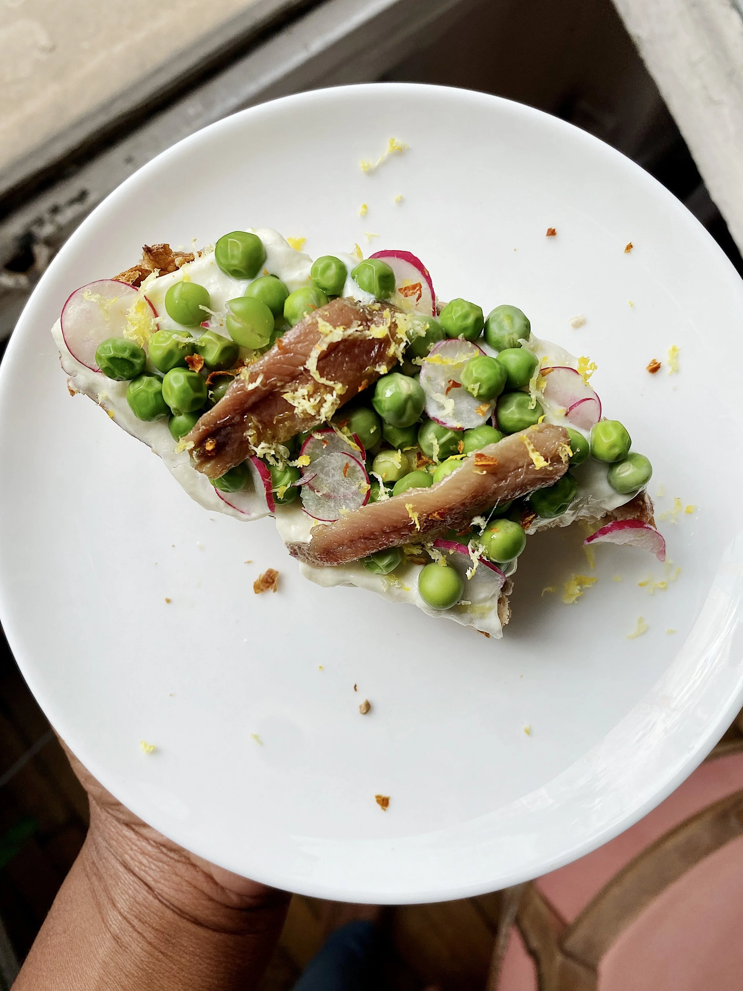 Hand holding a white plate with a slice of bread topped with green peas, radish slices, tuna, lemon zest, and some crumbs.