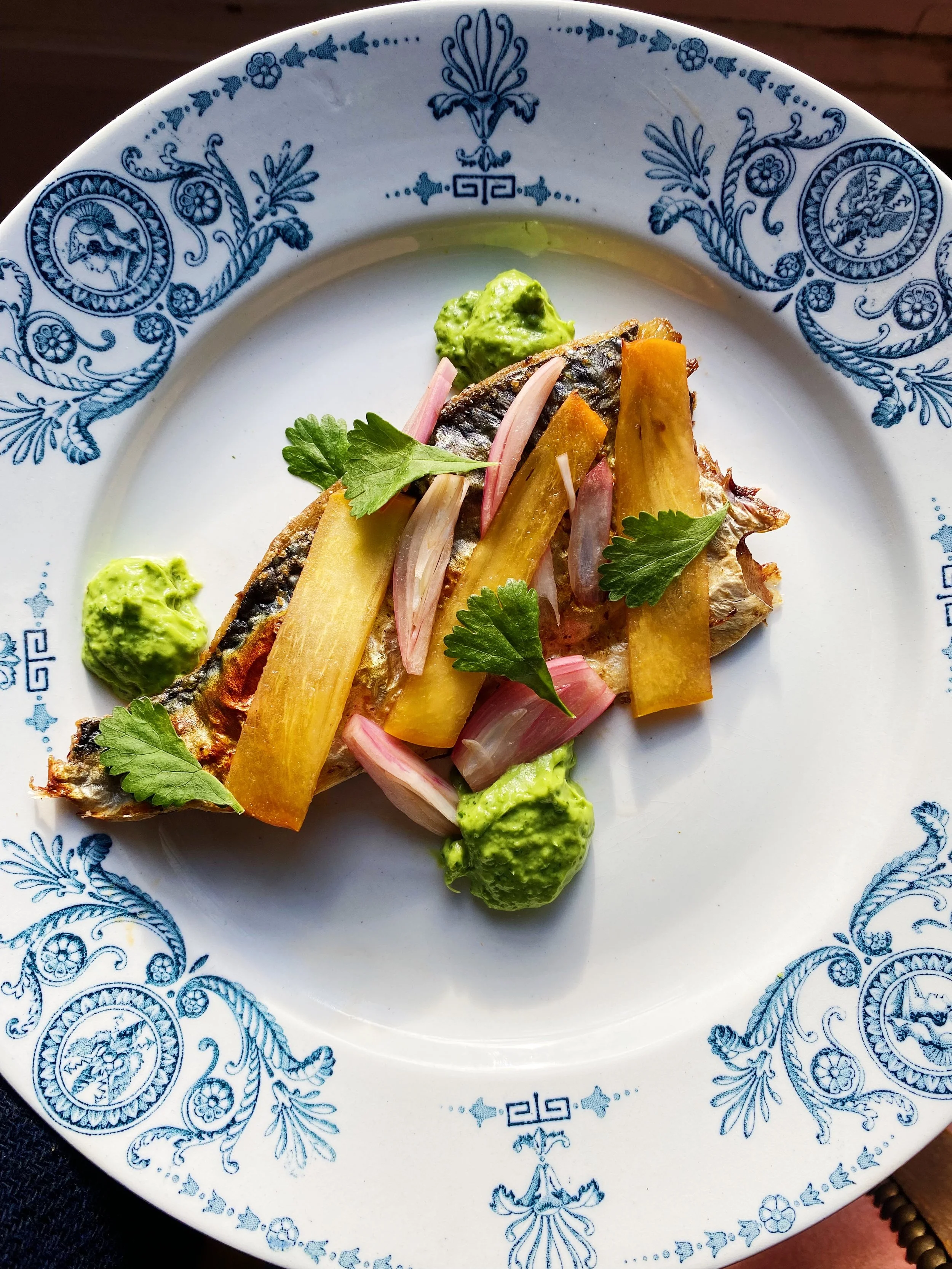 A white plate with intricate blue floral and geometric patterns around the rim contains a serving of grilled fish, garnished with fresh cilantro, thinly sliced pickled red onions, sliced yellow vegetables, and dollops of green sauce.