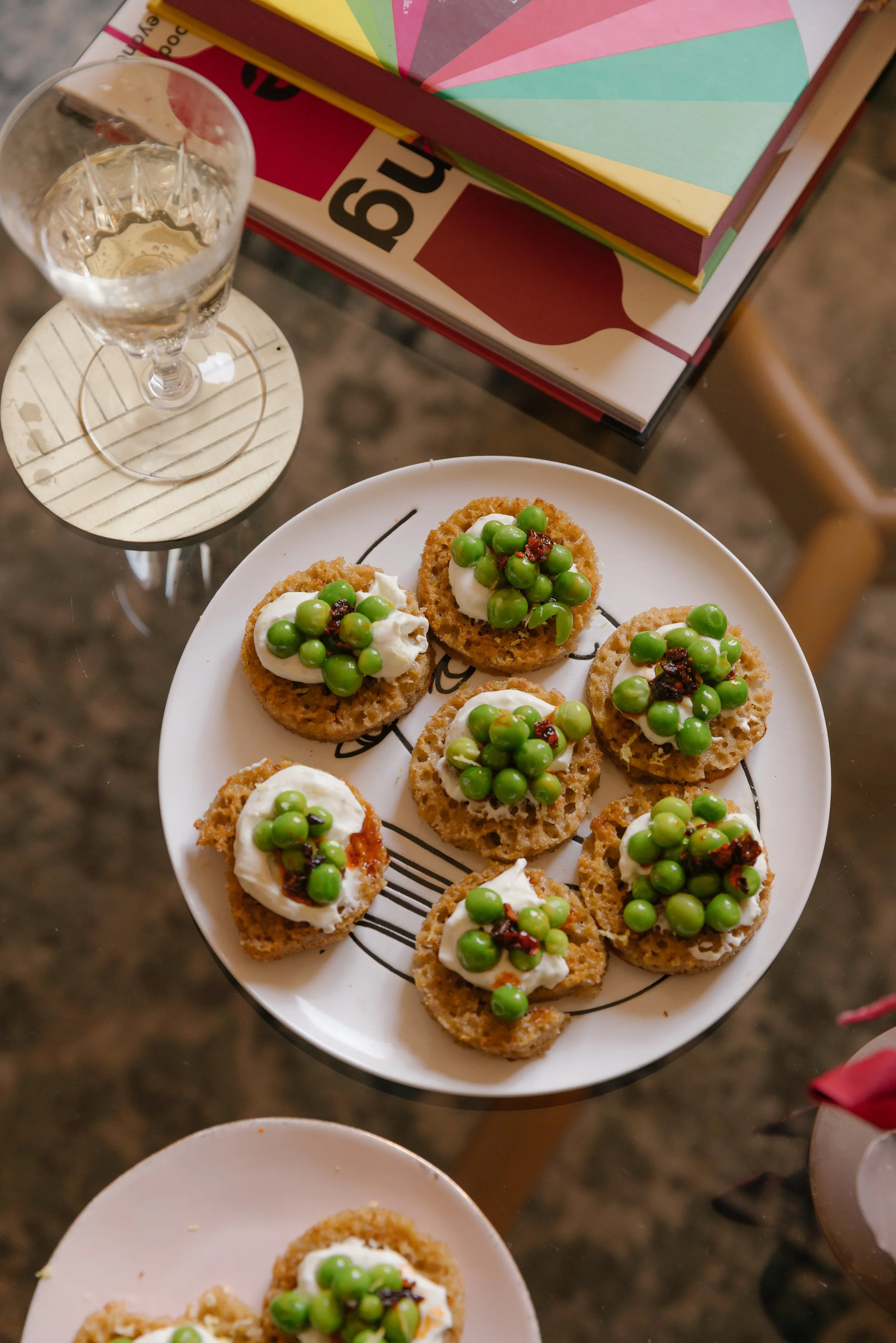 A white plate with several pieces of toast topped with cream cheese, green peas, and dried red peppers. A glass of water on a small wooden table, with colorful books and magazines nearby.