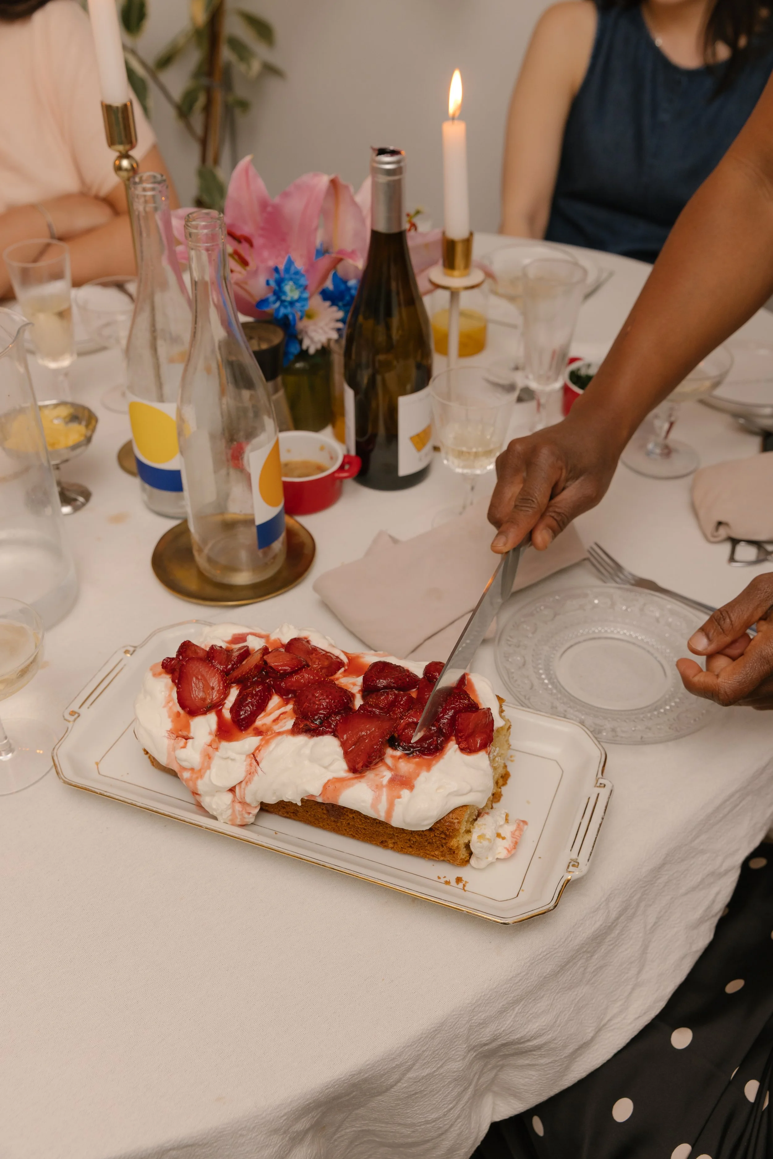 A dessert with whipped cream and strawberries on a rectangular cake on a decorative plate. The table has various glasses, bottles, and a floral centerpiece. People are gathered around, one person slicing the dessert.