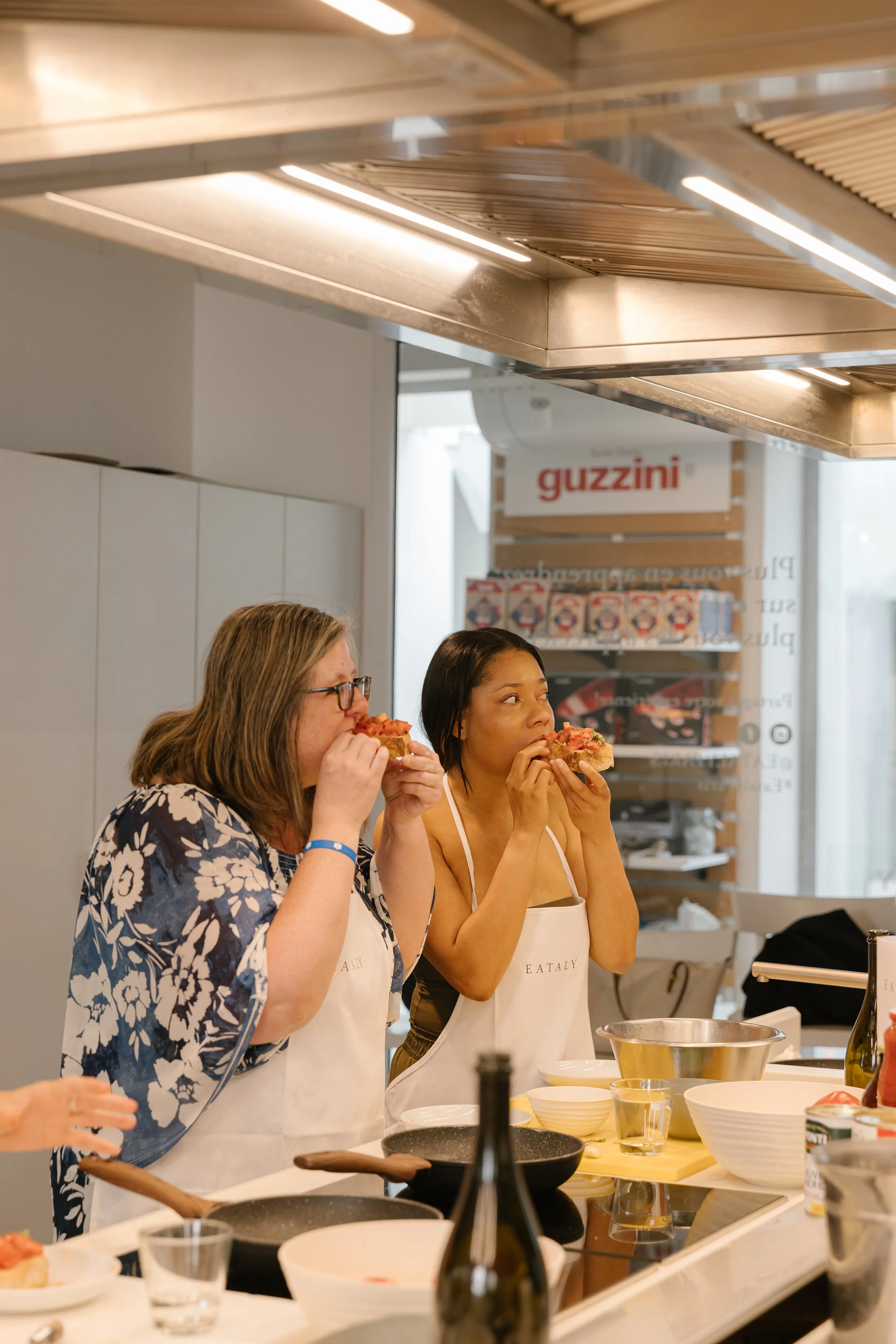 Two women wearing aprons tasting pizza in a modern kitchen with cooking utensils and ingredients on the counter.