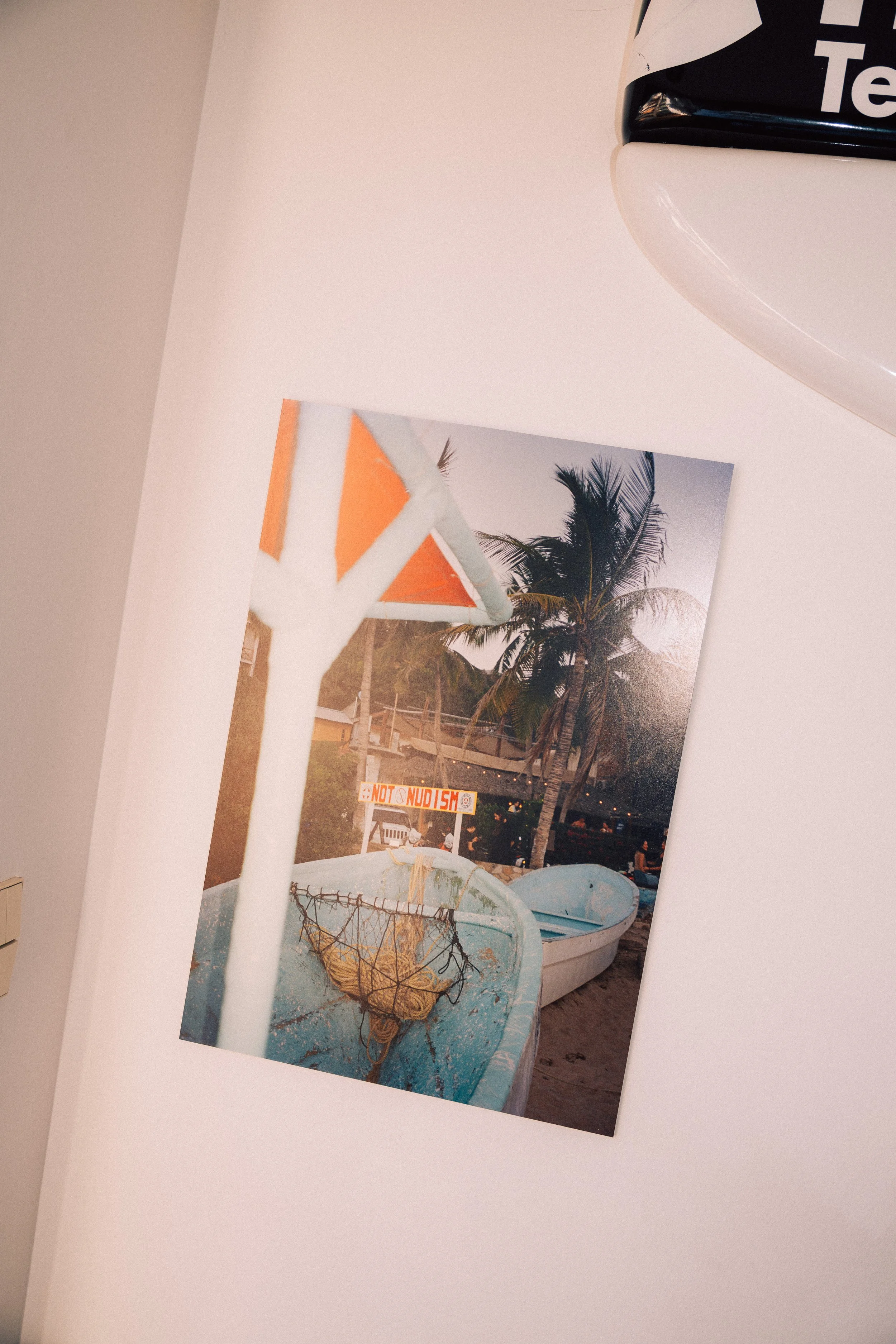 A printed photo on a white surface depicting a tropical scene with palm trees, boats, and a sign that reads 'NO NUDISM' with some boats docked near a shoreline at sunset.