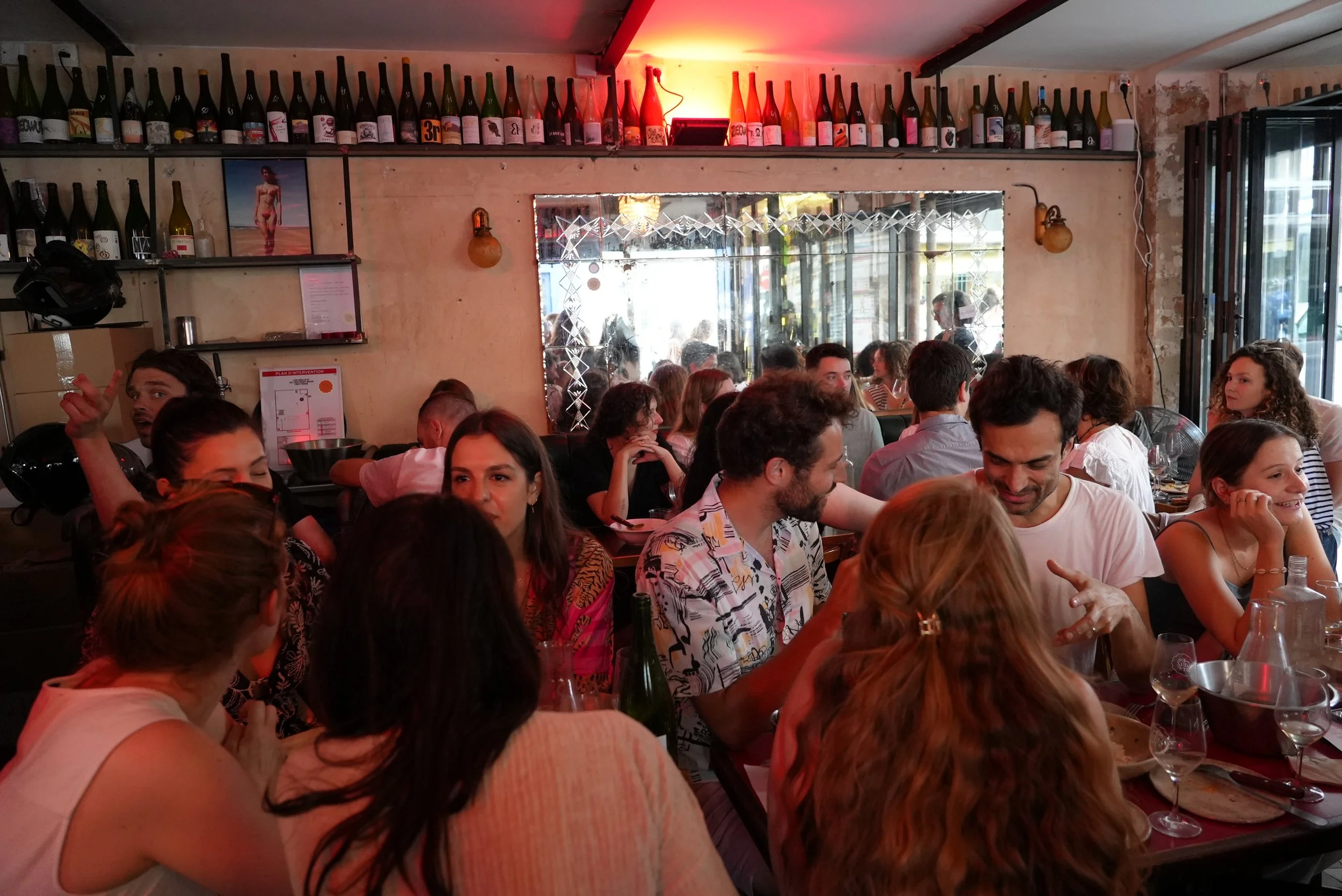 Crowded restaurant with people sitting at tables, bottles decorating the wall, a mirror reflecting the interior, and warm lighting.