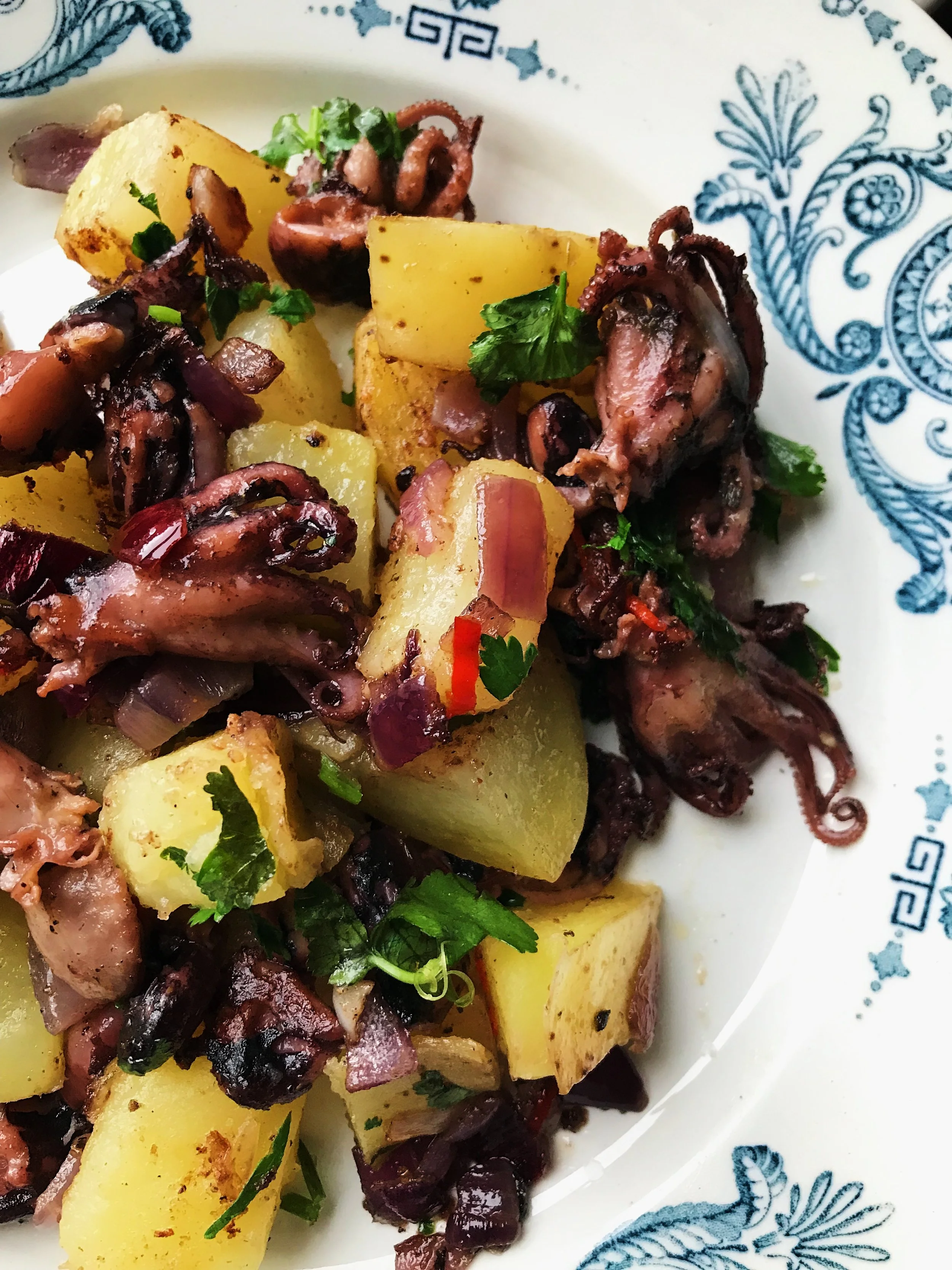 Grilled octopus with roasted potatoes and herbs on a decorative white and blue plate.